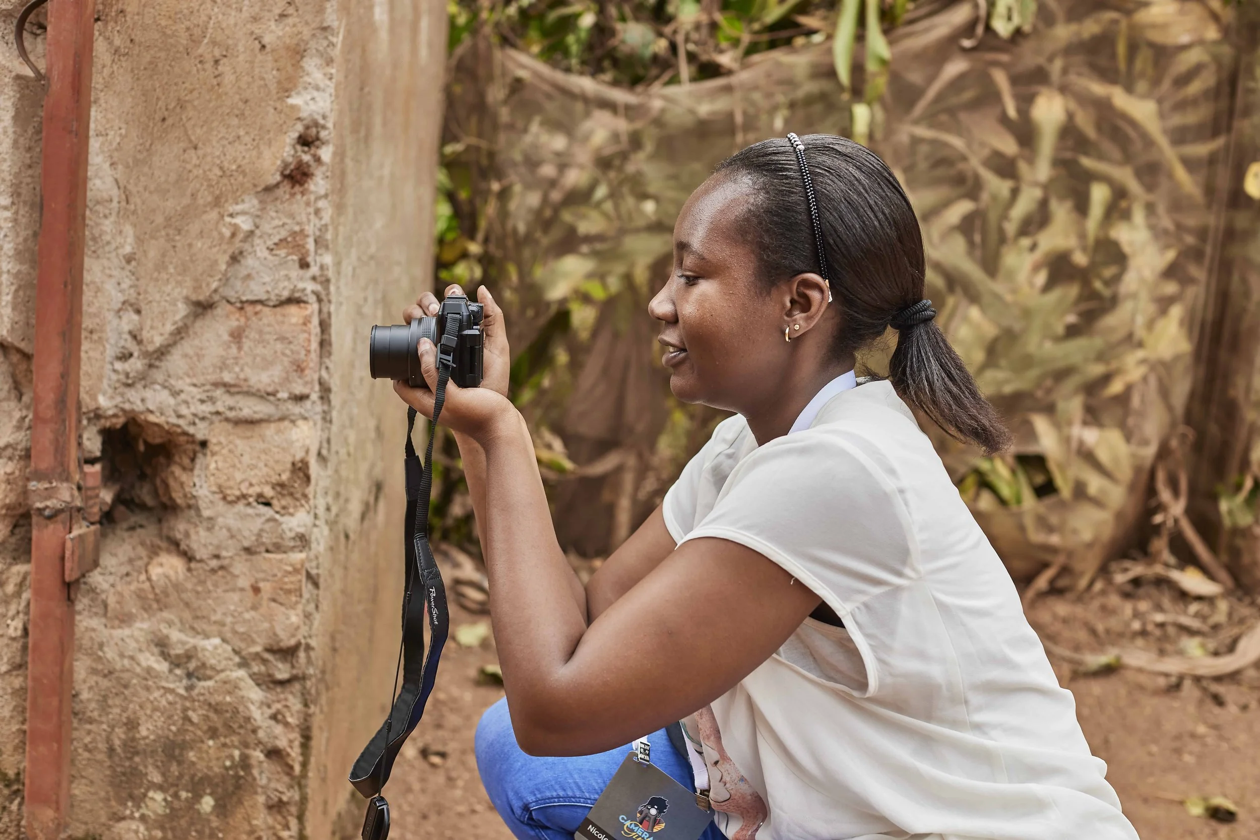A woman with dark hair tied back, wearing a white shirt, kneeling outdoors, taking a photograph with a camera aimed at a brick wall.