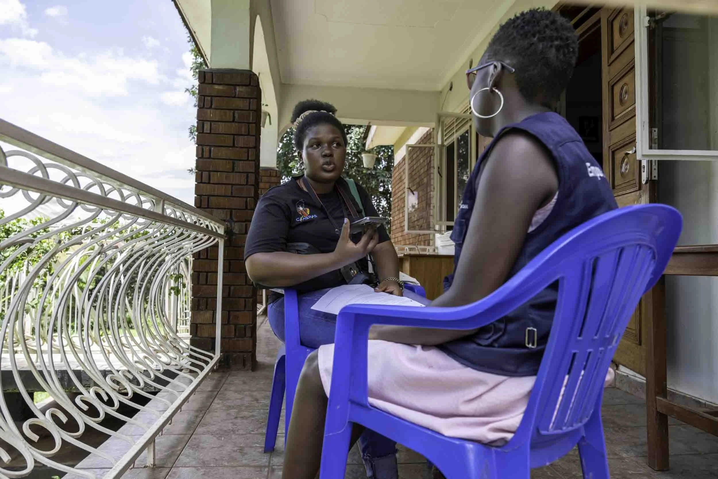 Two women having a conversation on a porch, sitting on blue plastic chairs. One woman, wearing glasses and a sleeveless shirt, is facing away from the camera. The other woman, wearing a black t-shirt, is holding a smartphone and looking at the woman 