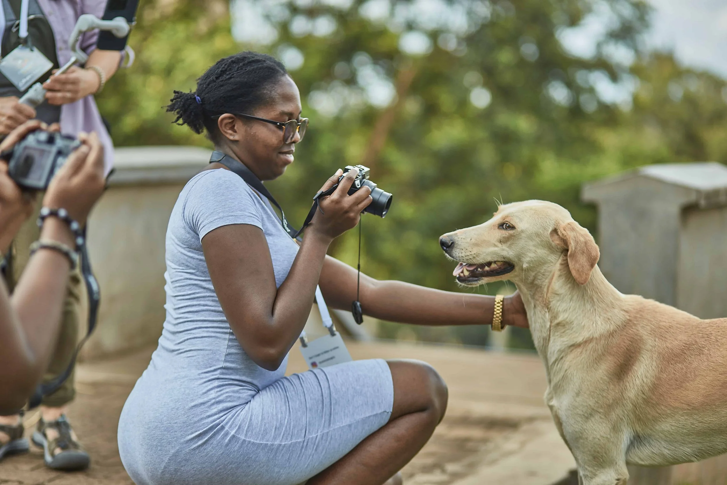 A woman with glasses and a gray shirt kneeling and smiling while holding a yellow Labrador Retriever at an outdoor event, with other people in the background.