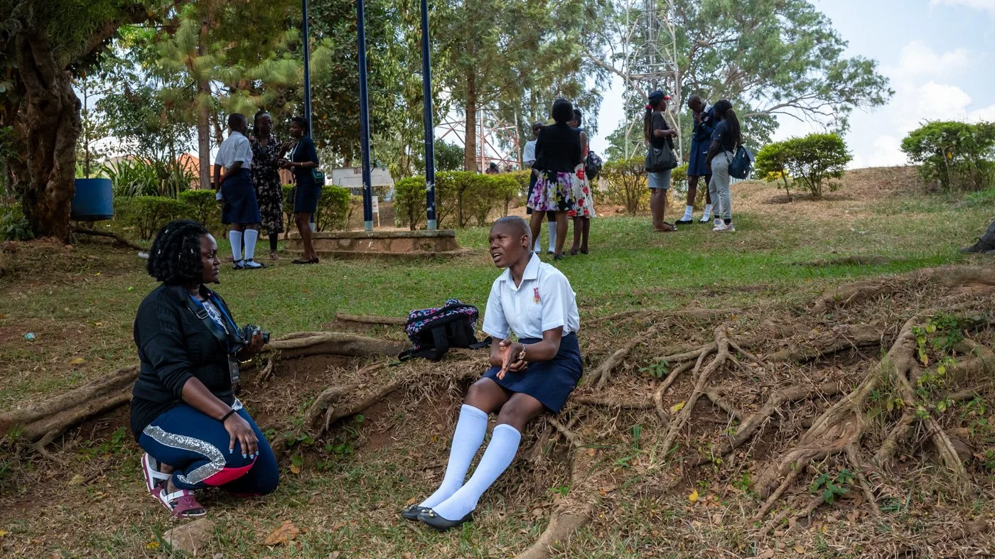 A schoolgirl in uniform sitting outdoors on a patch of grass, talking to a woman who is kneeling beside her, with a group of students standing and chatting in the background under a tree.
