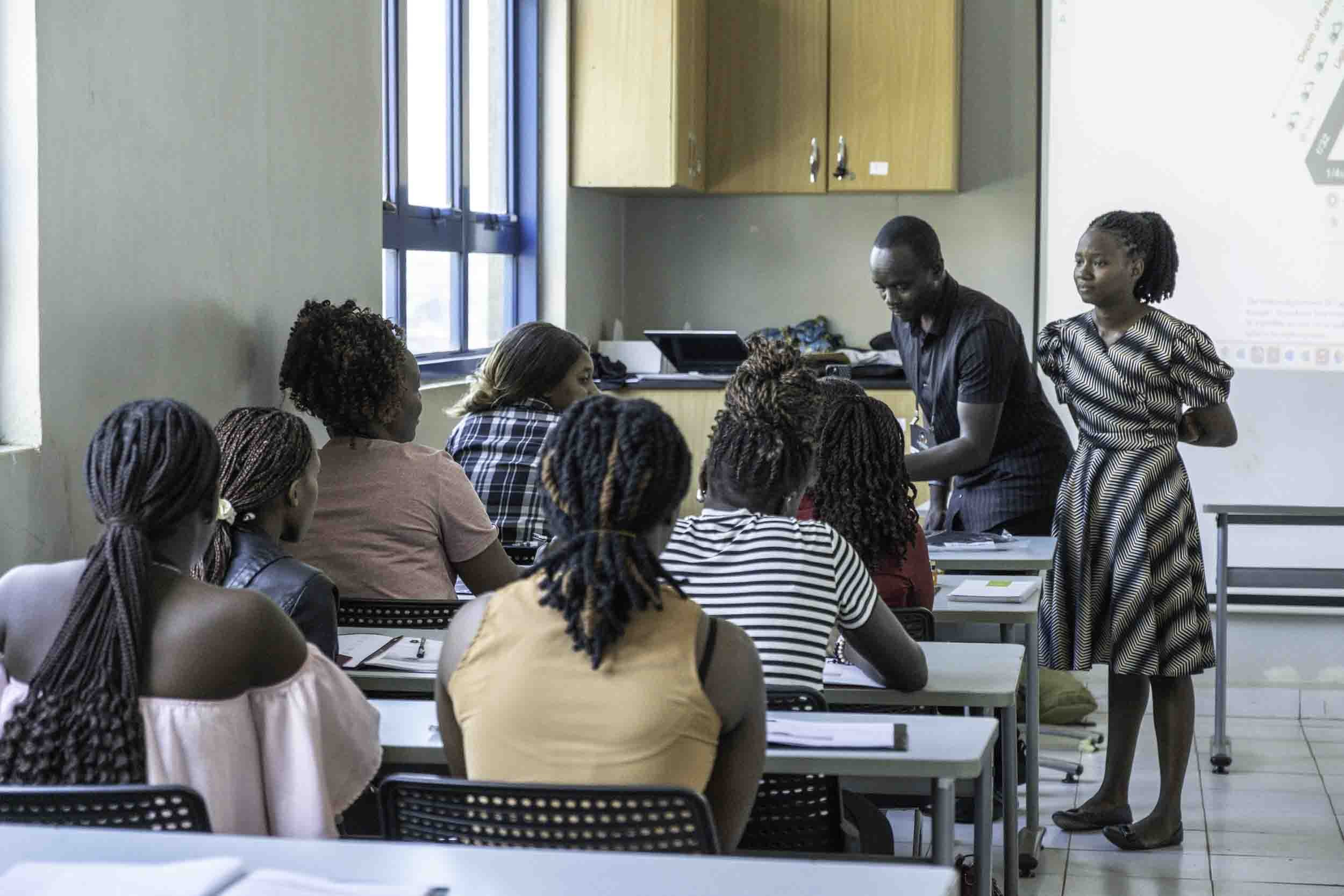 A classroom with students sitting at desks and listening to a woman standing near a whiteboard. The classroom has a window with blue frames and wooden cabinets. The woman appears to be teaching or presenting to the students.