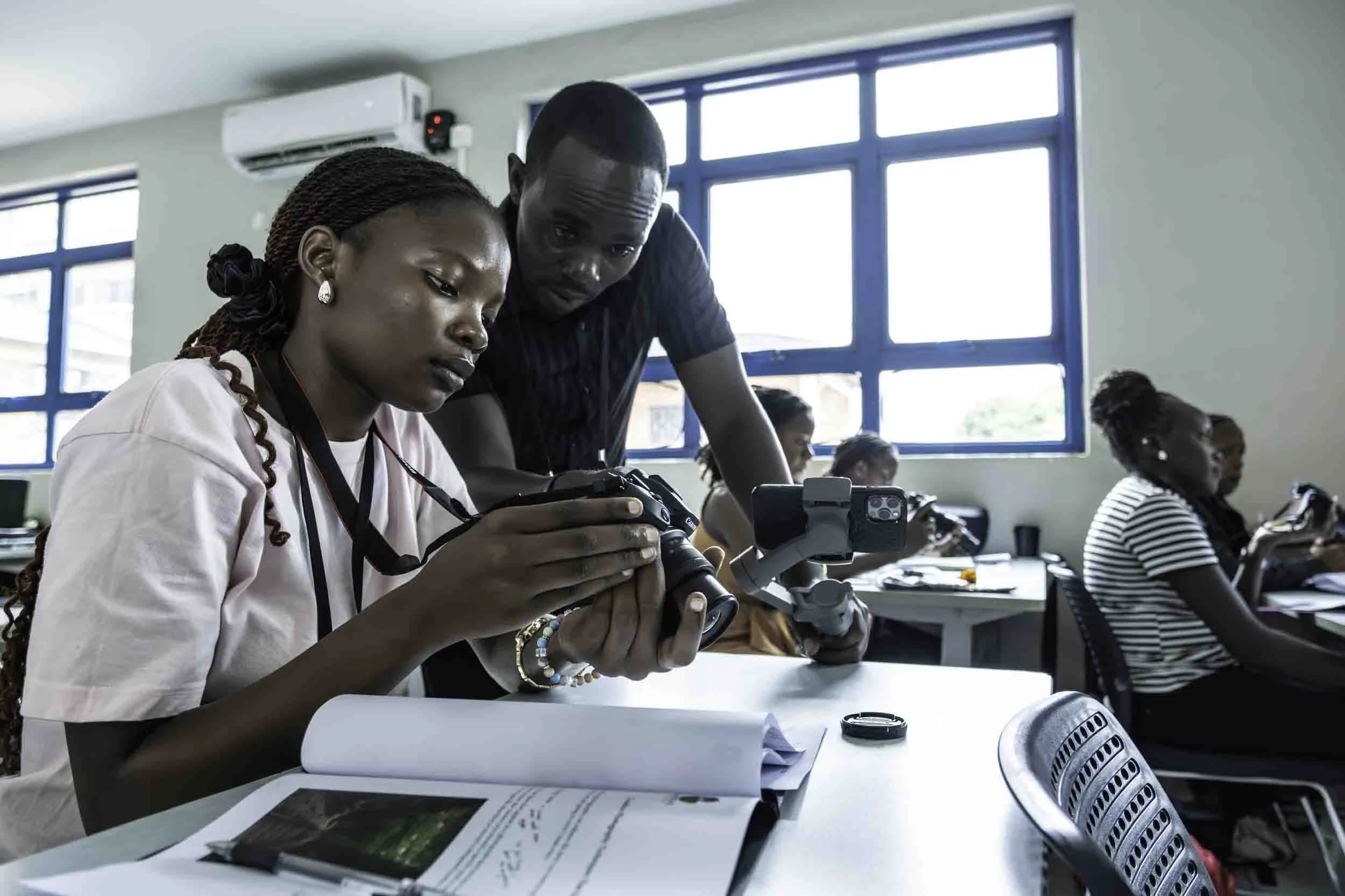 Two women examining a camera in a classroom, with other students seated at desks and large windows in the background.