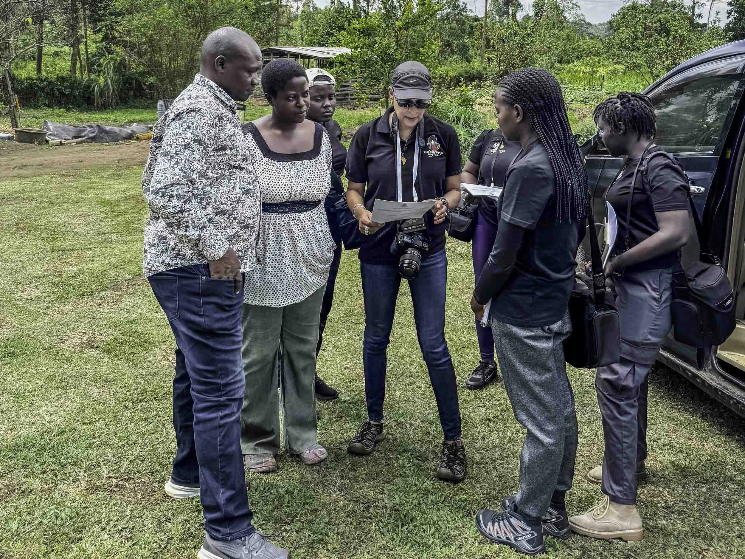 Group of six people gathered outdoors, closely examining documents and a camera, near a black vehicle.