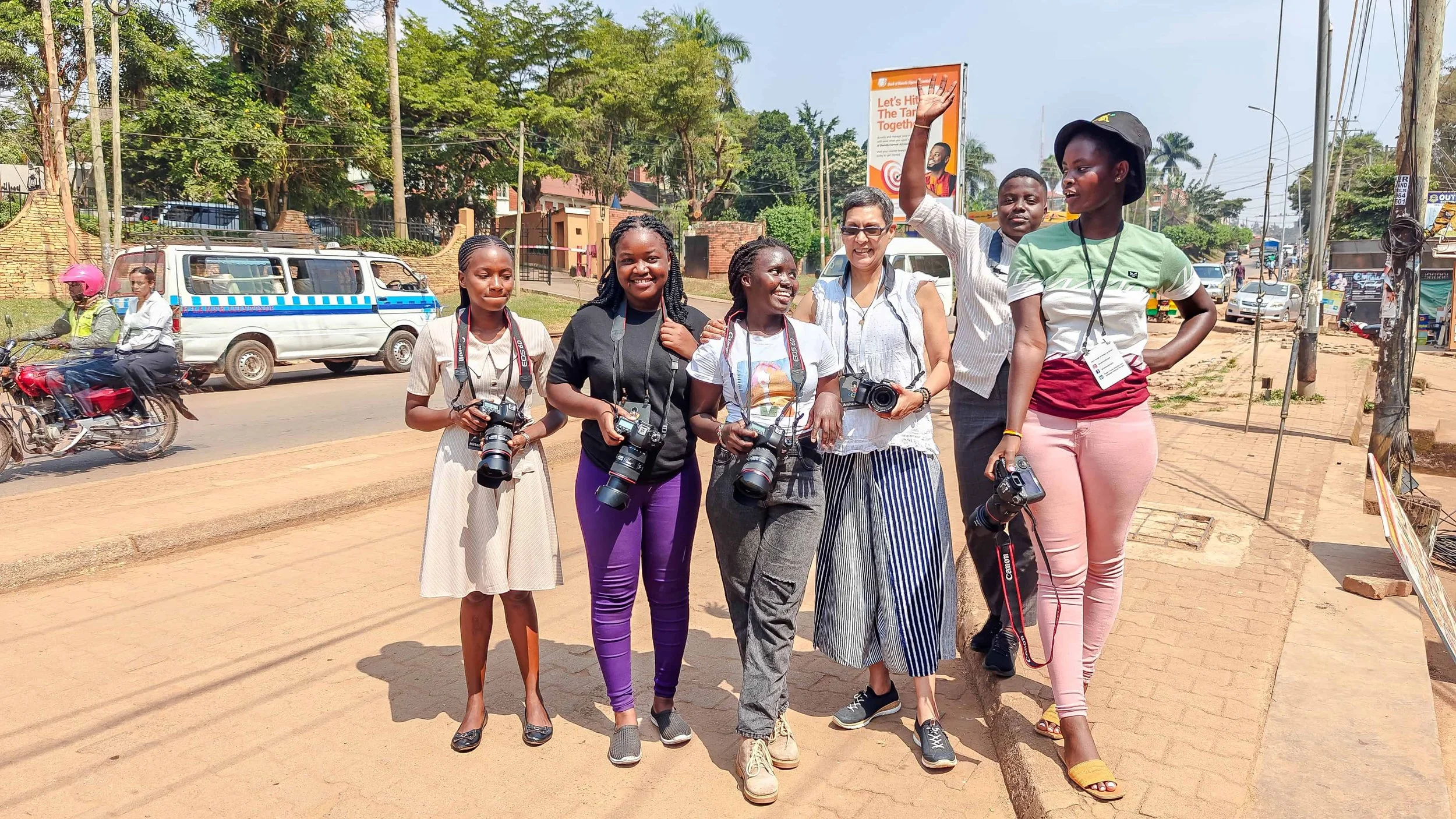 Group of five women with cameras and a man, smiling and standing together on a city sidewalk during daytime. Backdrop includes trees, vehicles, and a billboard.