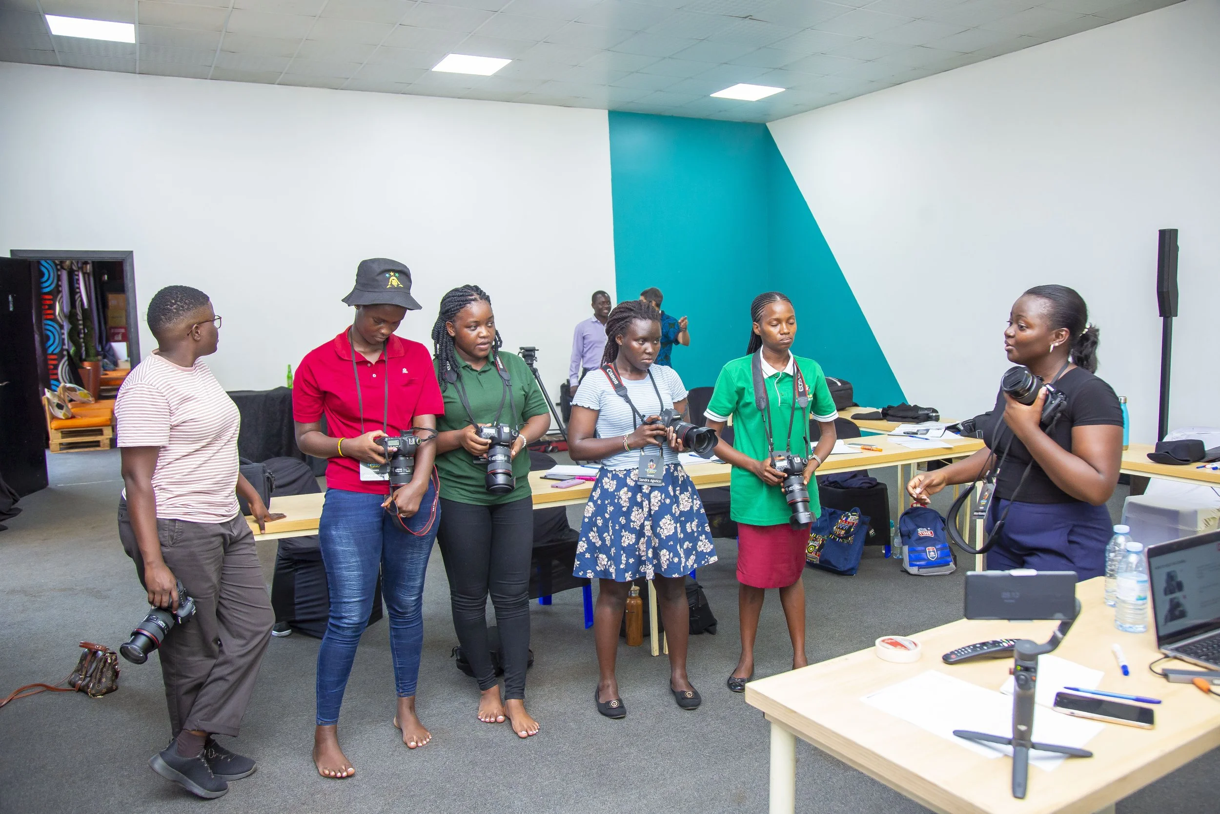 A group of women standing in a circle in an indoor space, some holding cameras, with a woman speaking to them.