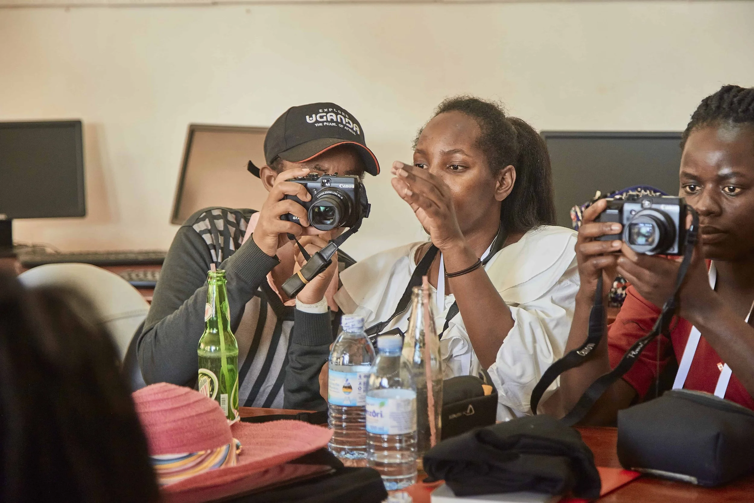 Three women sitting at a table with various bottles and papers, one woman is taking a photo with a camera, and a woman is whispering to her.