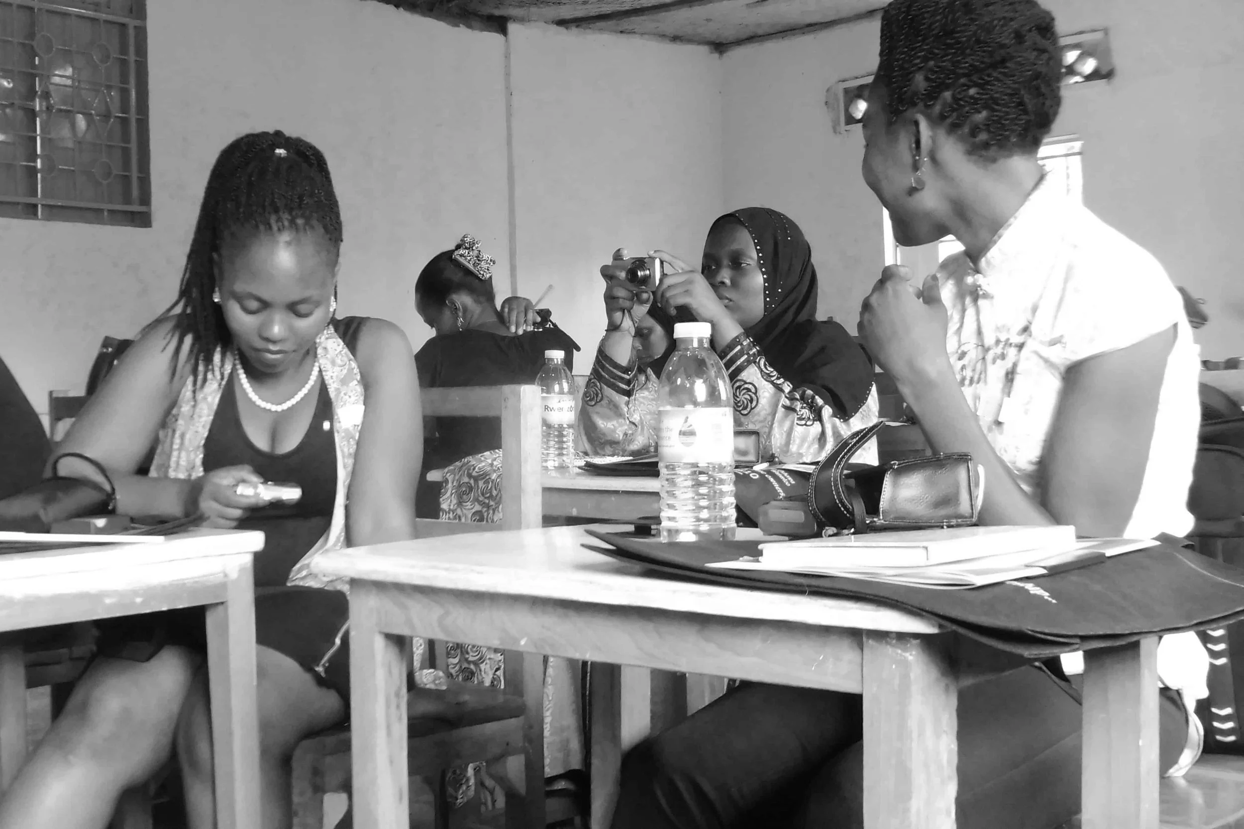 Four women sit at a table in a room with plain walls, engaged in various activities. One woman in the foreground is looking at her phone, wearing a sleeveless dress with a pearl necklace. The second woman, in the middle, is holding a small camera, we