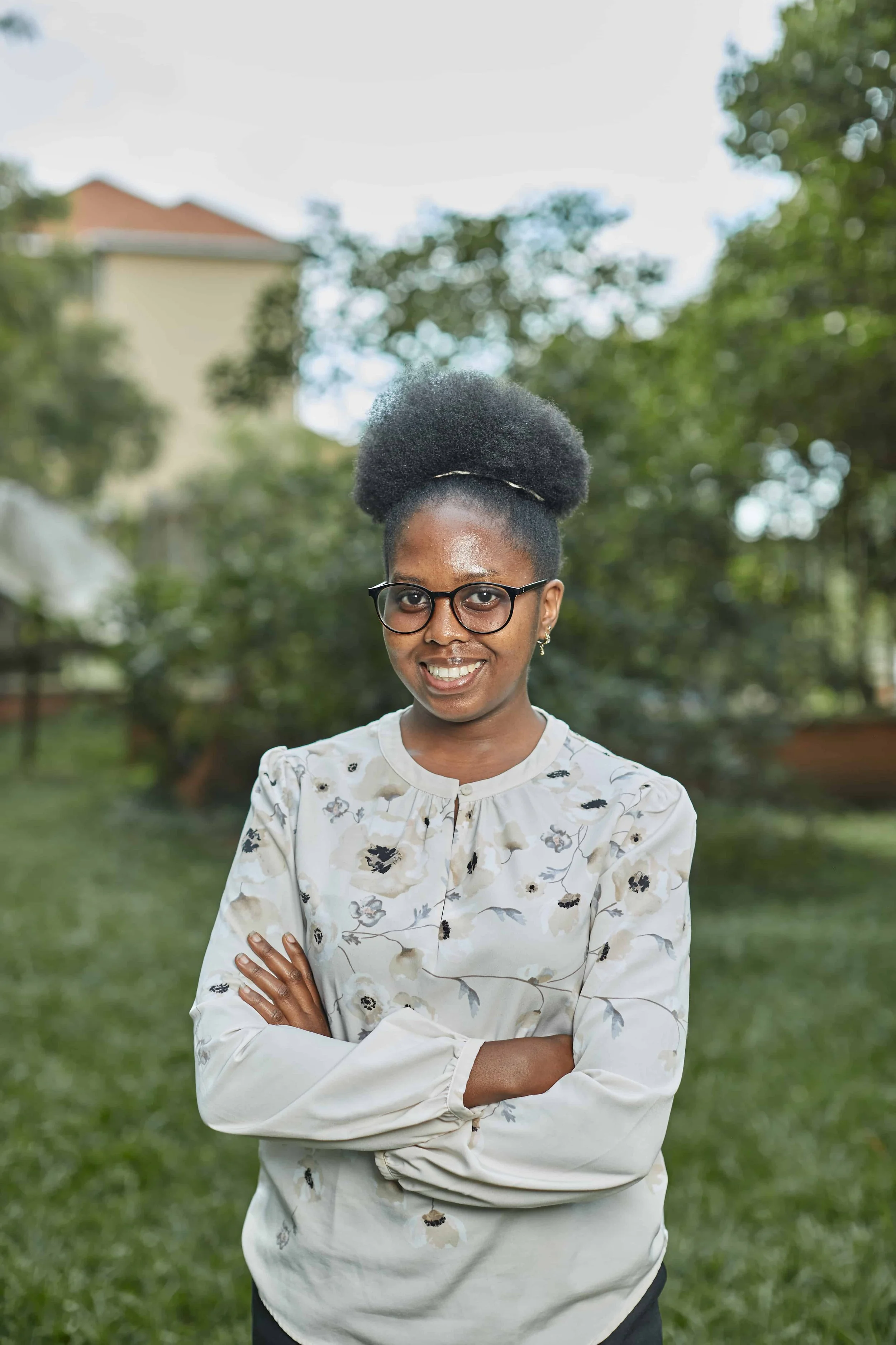 A woman with glasses and a natural hairstyle, wearing a long-sleeve beige blouse with a floral pattern, standing outdoors in a park with trees and a building in the background.