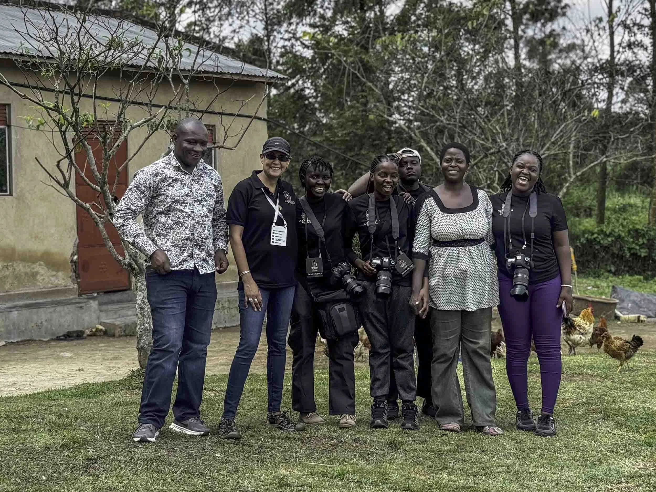 A group of people, including men and women, standing outdoors on grassy ground with chickens nearby. They are smiling and some have cameras hanging from their necks. In the background, there is a small building with trees and foliage.