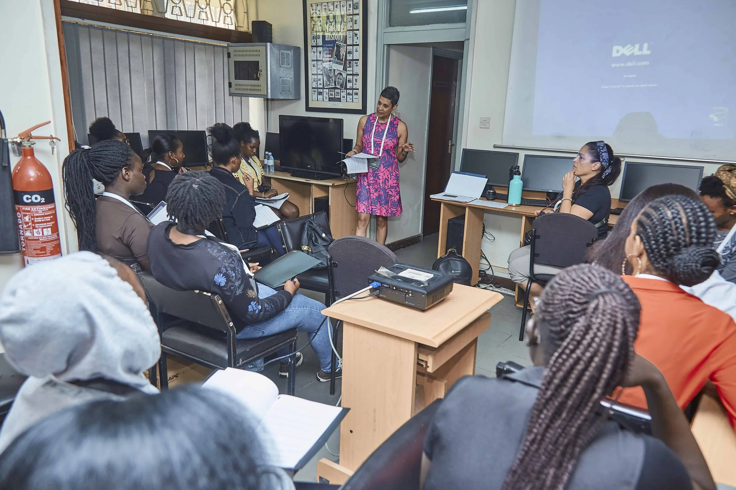 A group of students attending a classroom lecture with a woman teacher. The teacher is standing near a wall, holding papers, and speaking to the students, some of whom are taking notes. The classroom contains computers, a projector, and posters on th