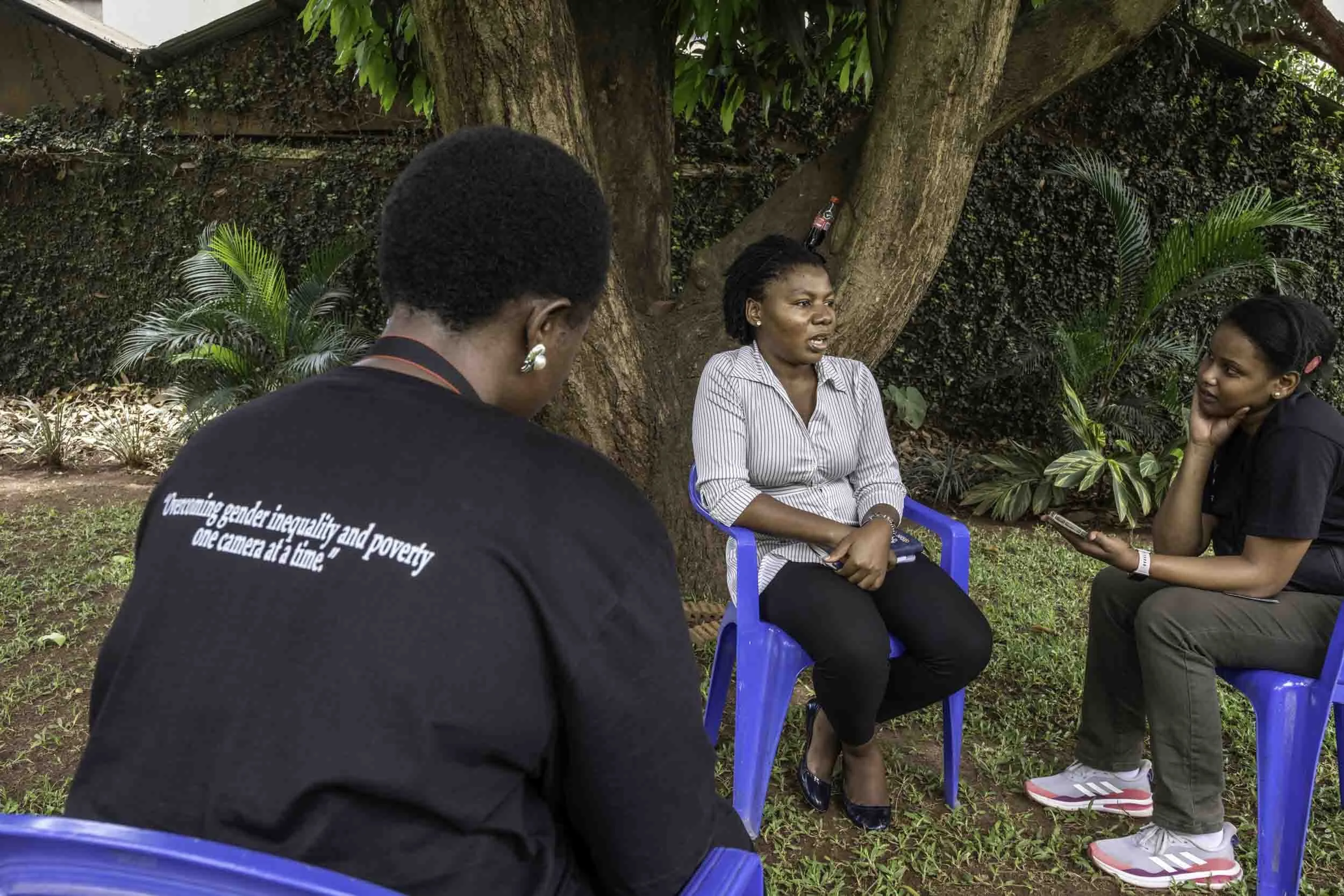 Three women engaged in conversation outdoors, sitting on blue plastic chairs near a large tree with green foliage, in a garden setting.