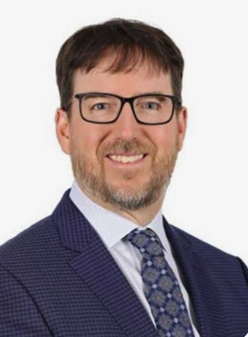 Professional headshot of a man with short brown hair, beard, wearing a dark blazer, light sweater, and collared shirt against a neutral background.