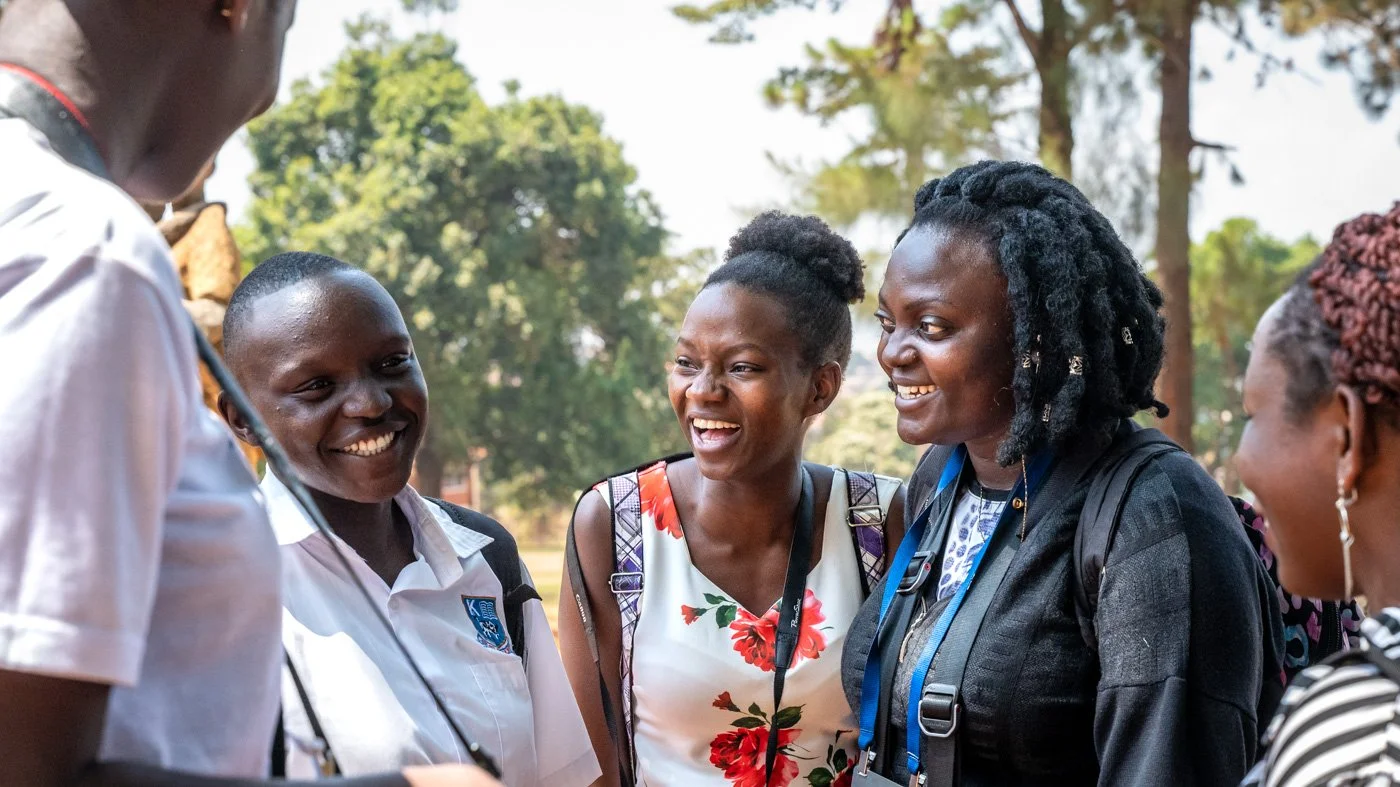 Group of women smiling and talking outdoors in a park with trees in the background.