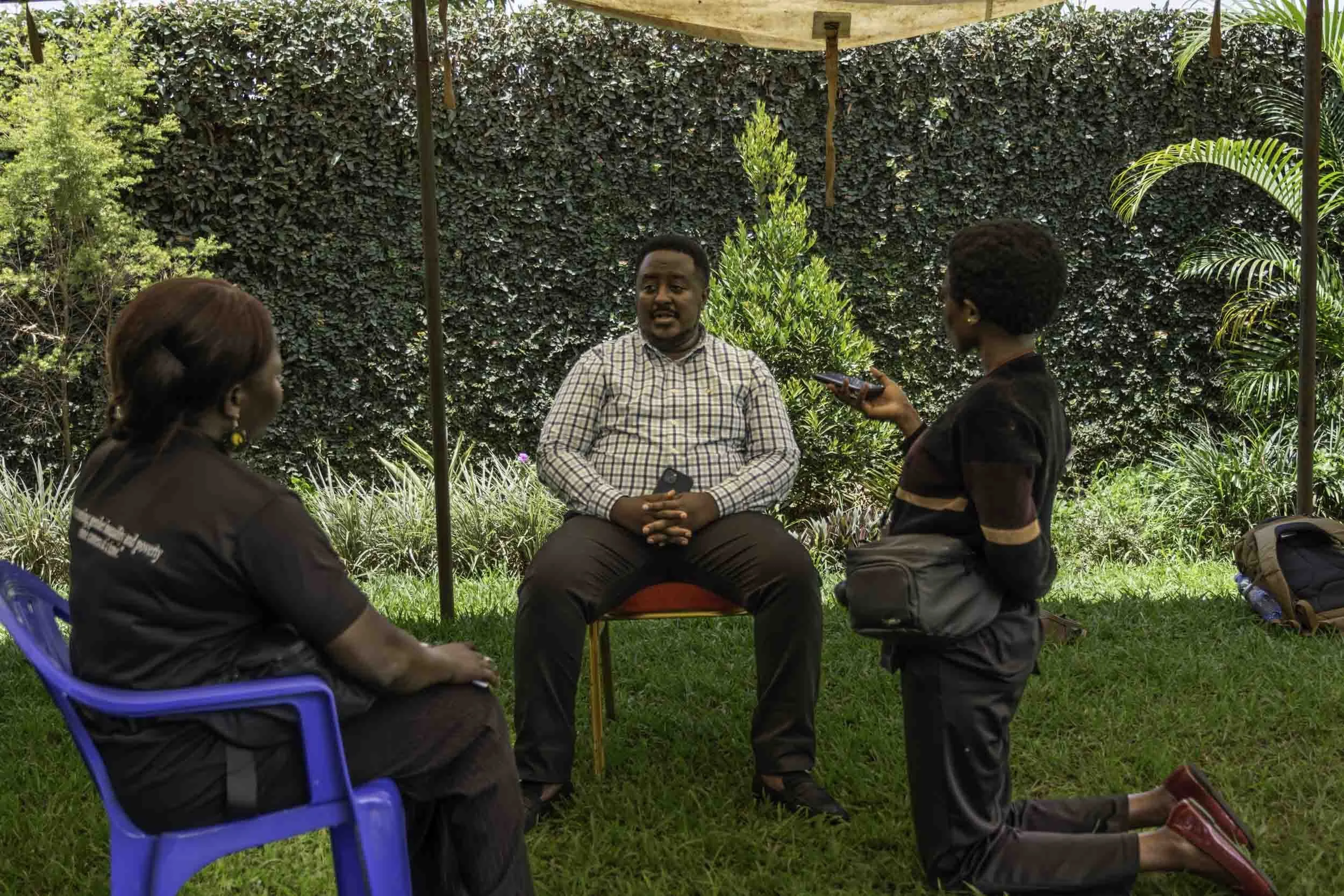 A man and two women sitting outdoors on grass, engaging in conversation, with lush green plants and a hedge in the background.