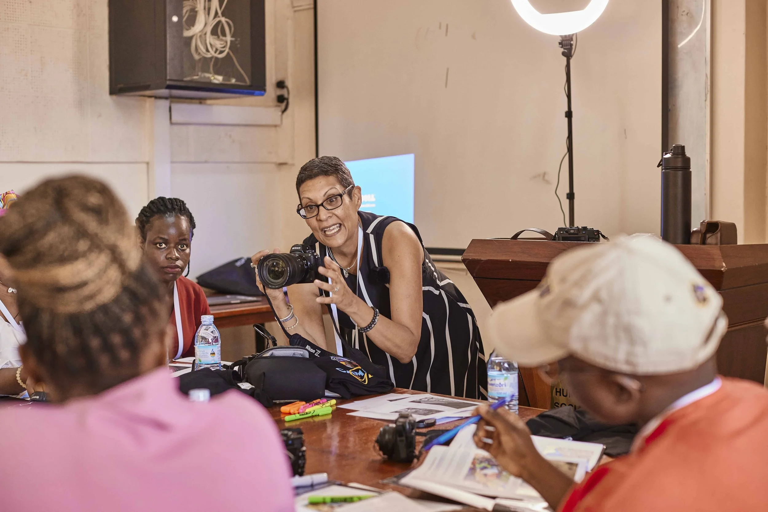 A woman with short hair and glasses holding a camera and talking to a group of women seated around a table with papers, water bottles, and writing supplies, in a classroom or workshop setting with whiteboard and projector in the background.