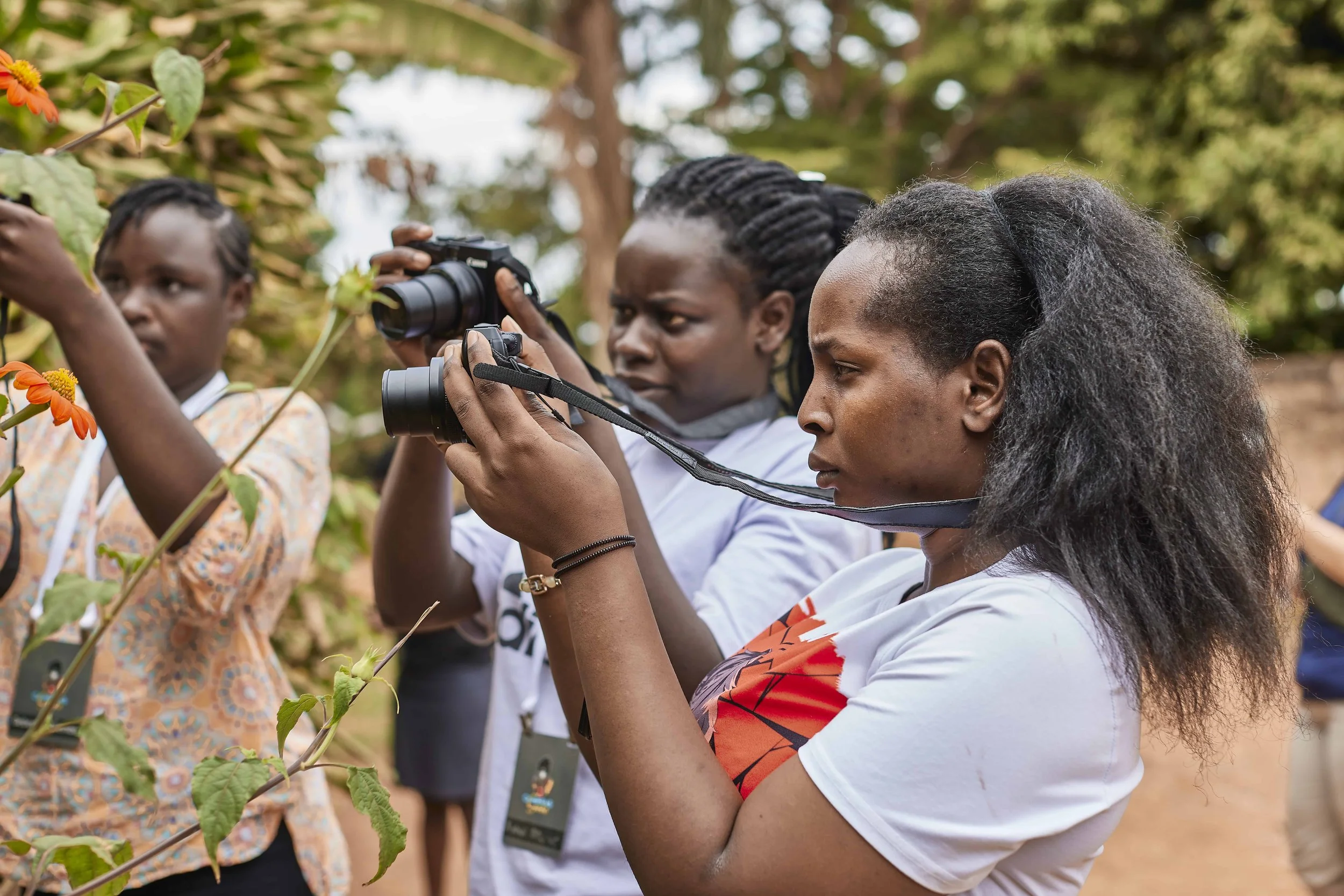 Three women with dark skin and black hair, two with braids and one with curly hair, observing plants outdoors and one woman taking photos of the plants with a camera.