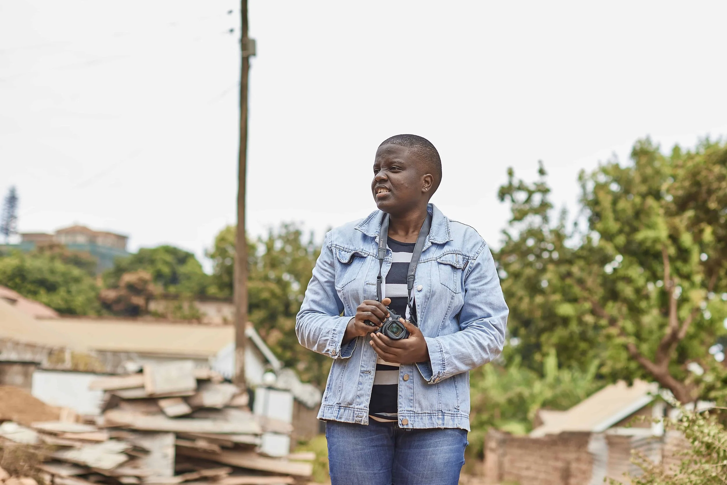A young woman holding a camera outdoors, wearing a denim jacket and a striped shirt, with trees and buildings in the background.