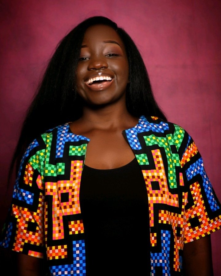 Portrait of a smiling Black woman with long black hair, wearing a vibrant, multicolored geometric Pattern jacket over a black top, standing against a pink background.