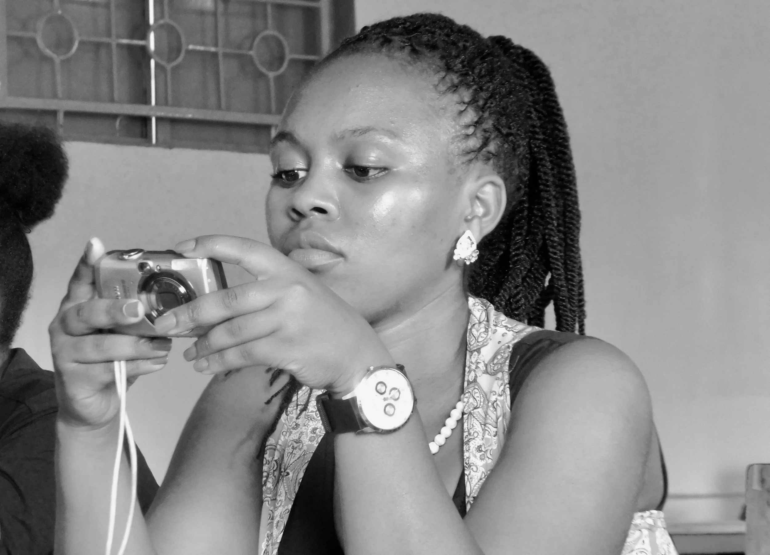 A woman with braided hair, wearing earrings and a patterned sleeveless top, taking a selfie while having her blood drawn, with a blood pressure cuff on her arm and a medical device around her wrist.
