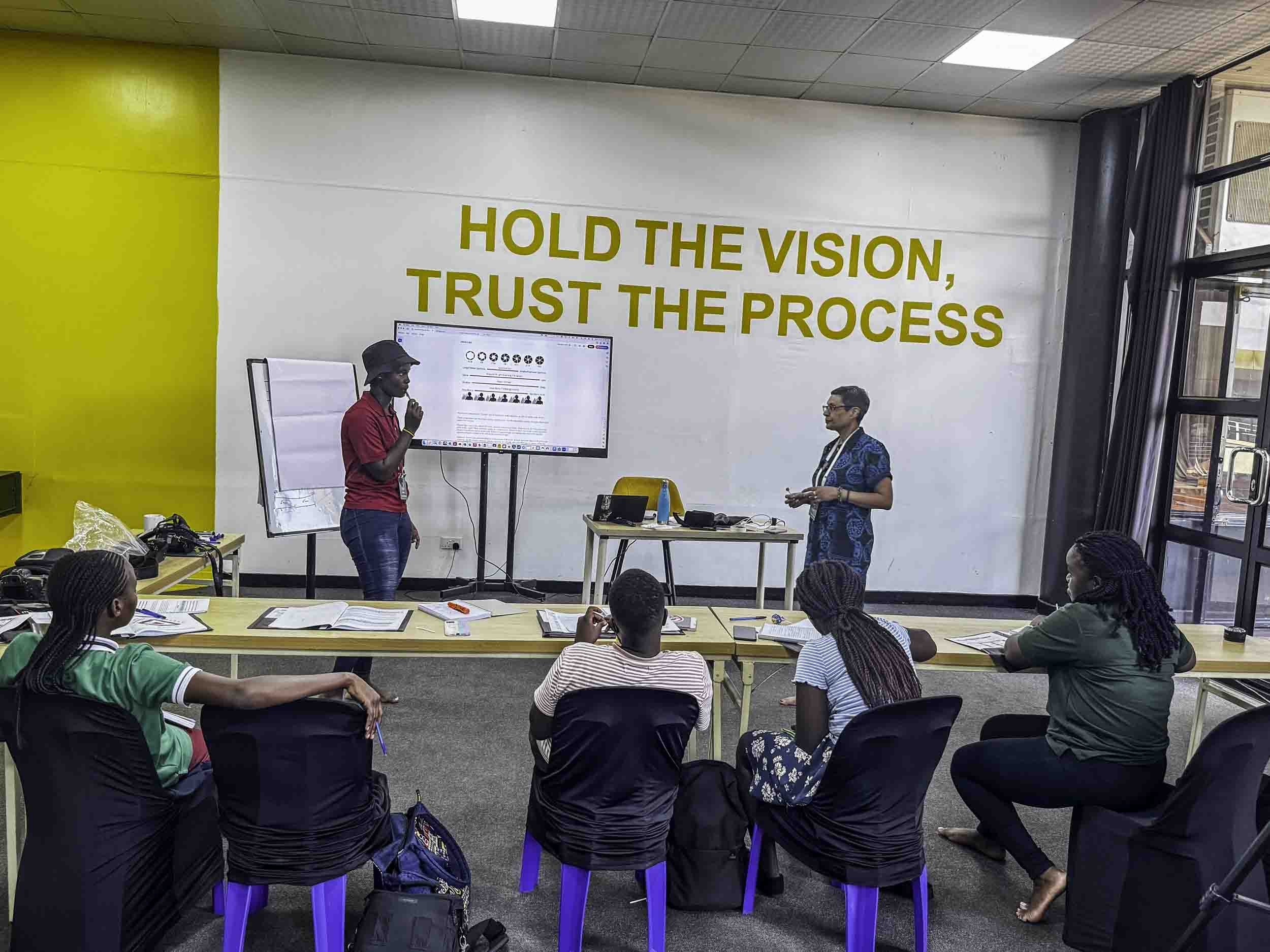 A classroom or conference room with a group of students or attendees seated at tables, listening to a presenter. The presenter, wearing a red shirt and black hat, is speaking into a microphone and standing near a large screen displaying a webpage. A 