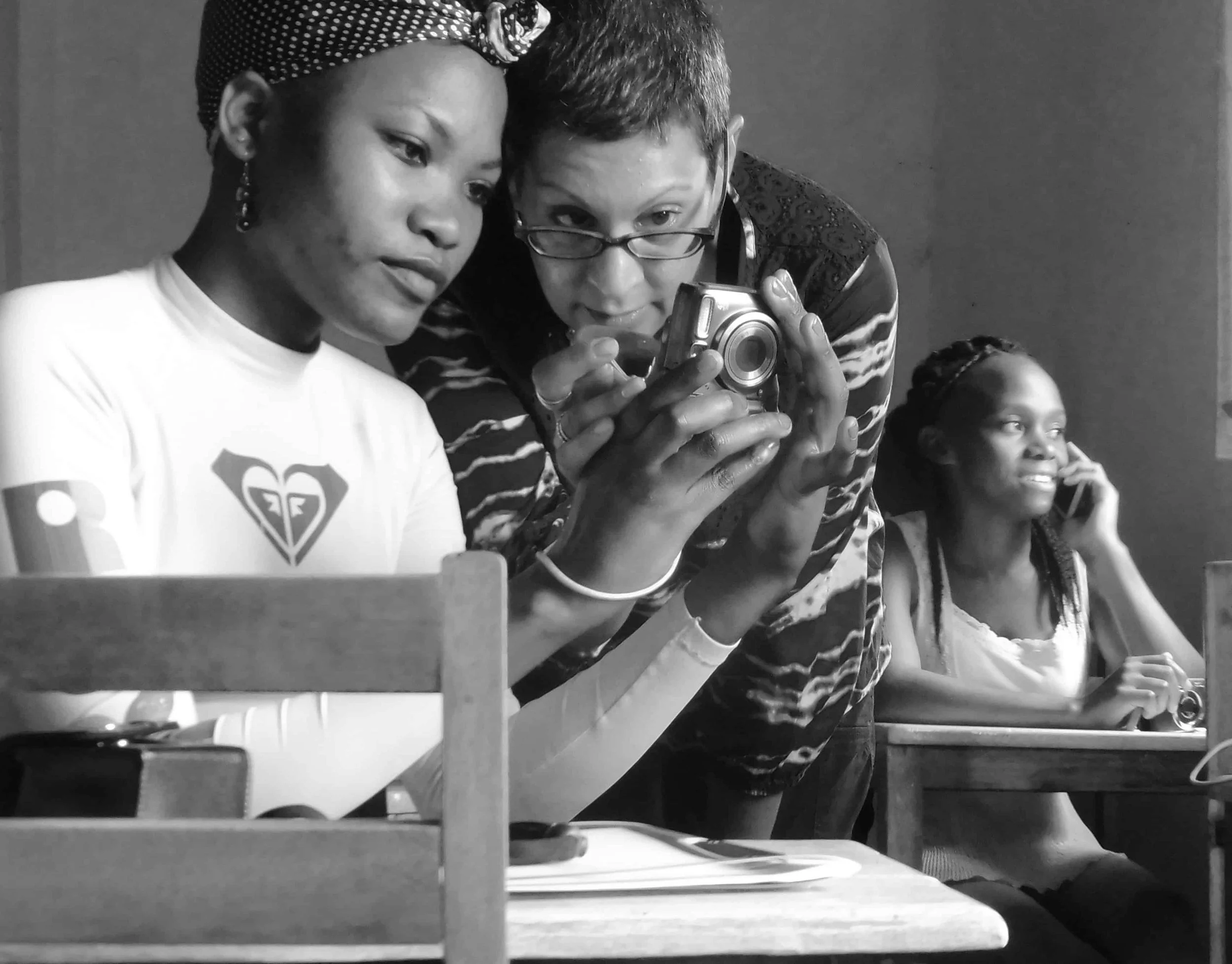 Two women sitting side by side at a table, one wearing glasses and reading a camera, the other in a white t-shirt with a heart-shaped design, both focused on something in front of them. They appear to be in a casual setting.