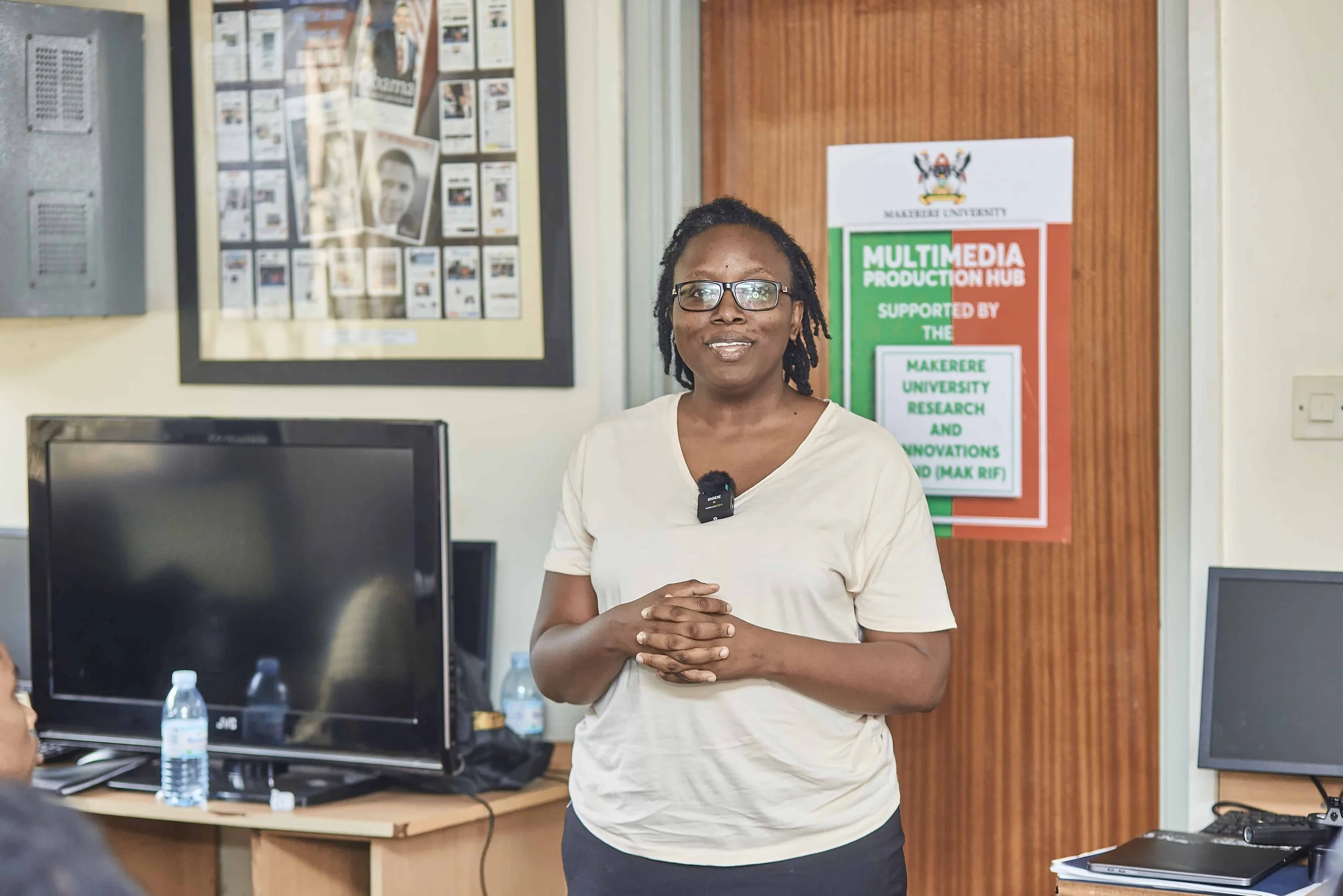 A woman with glasses and dreadlocks smiling in front of a poster at Makerere University Multimedia Production Hub.