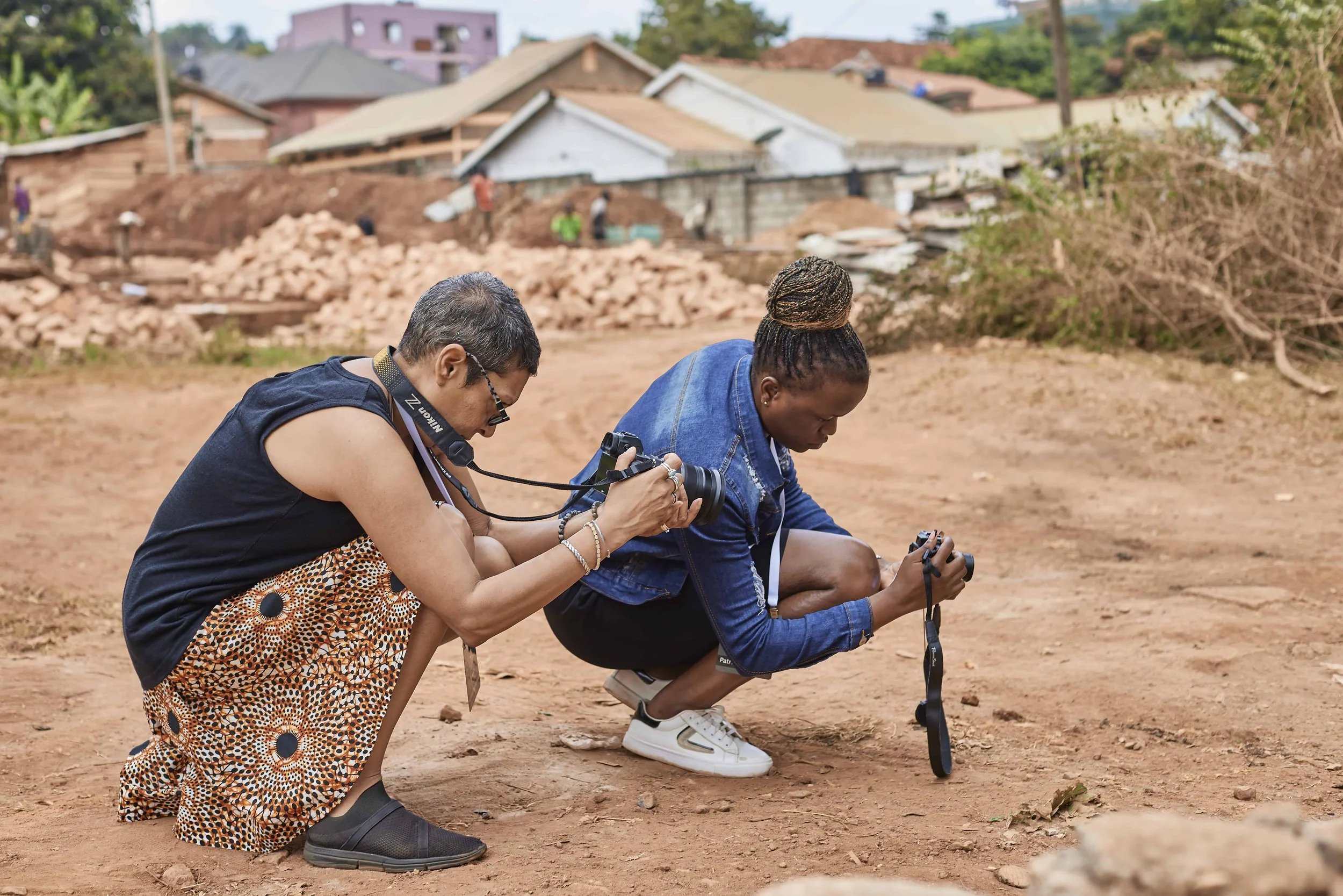 Two women kneeling on the ground, examining a metal detector in a dirt area with houses and trees in the background.