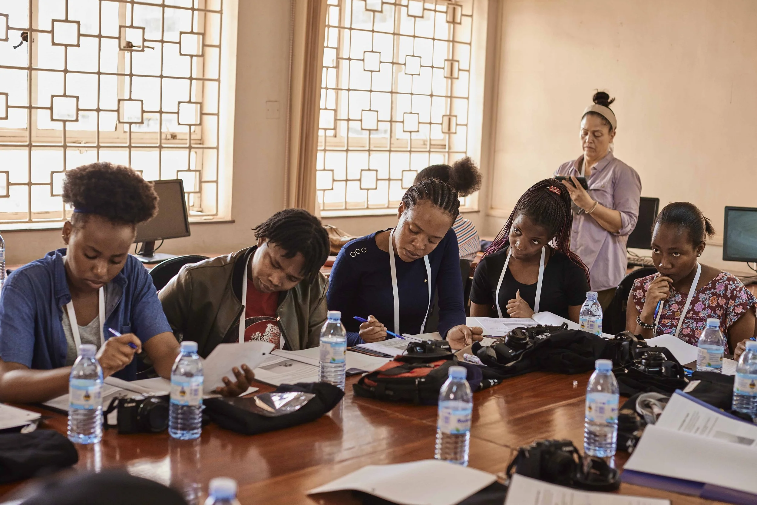 A group of diverse women sitting at a conference table with documents, pens, cameras, and bottled water, engaged in a workshop or training. In the background, a woman stands and uses her phone in a room with large windows and patterned window frames.