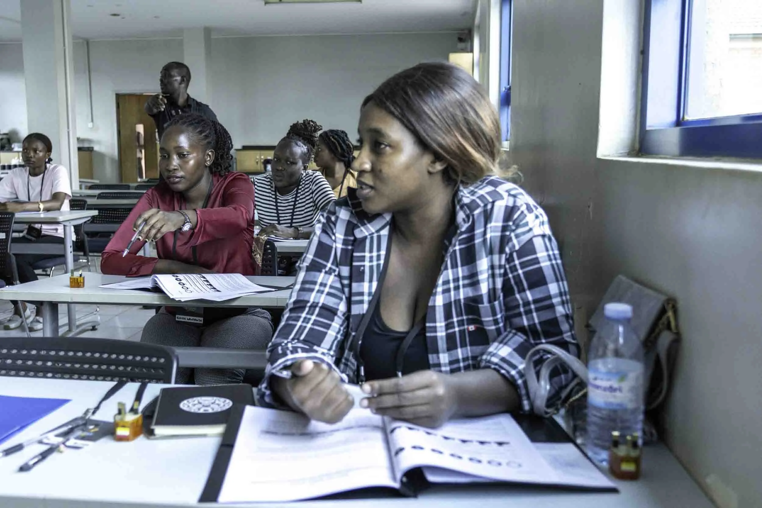 Several women seated at desks in a classroom or conference room, engaging in a discussion or lecture. A man is standing in the background, pointing. The women have various papers, notebooks, and a water bottle on their desks.