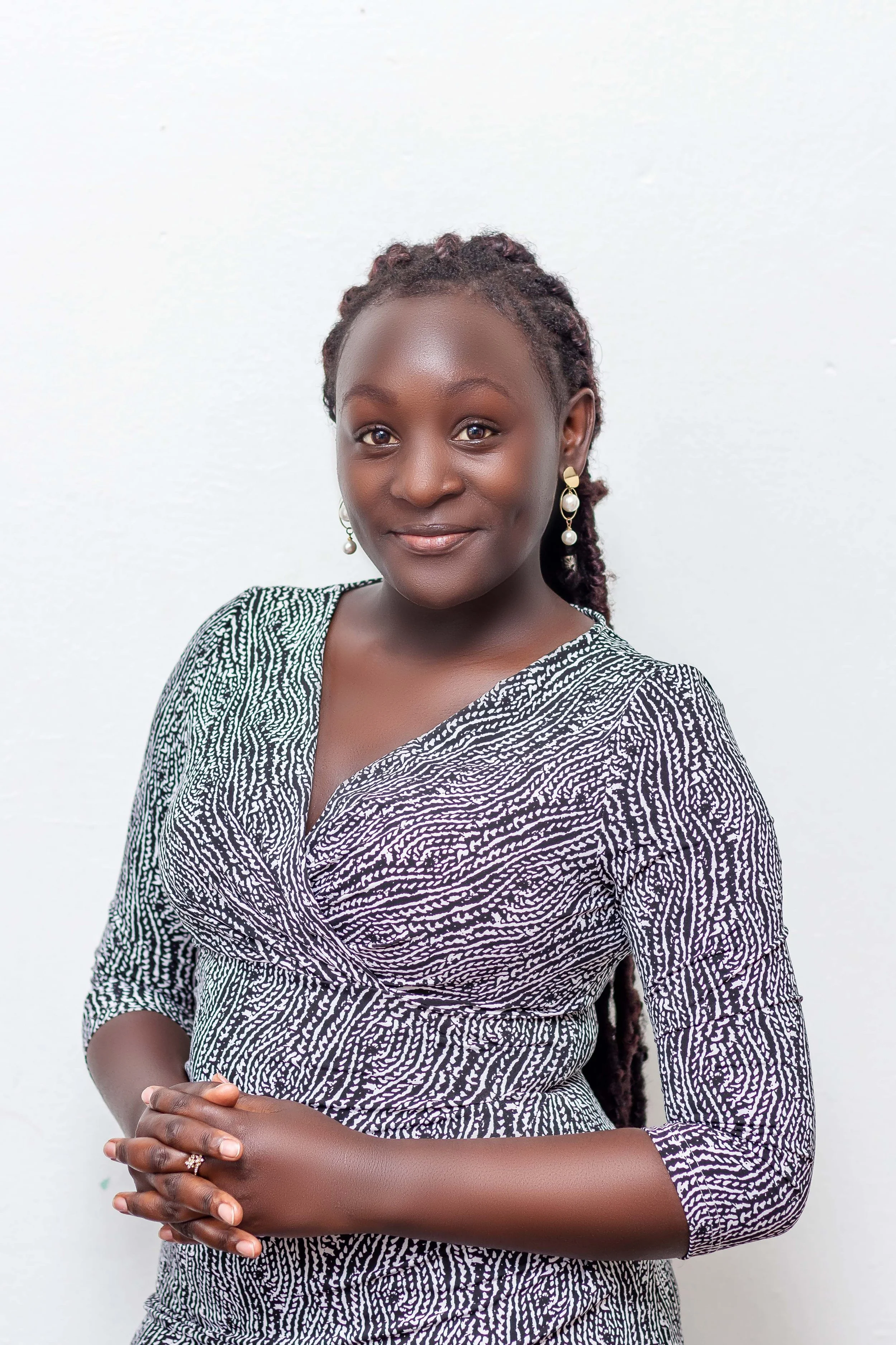 Portrait of a woman with dark skin, braided hair, and pearl earrings wearing a black and white patterned dress, standing against a plain white background.