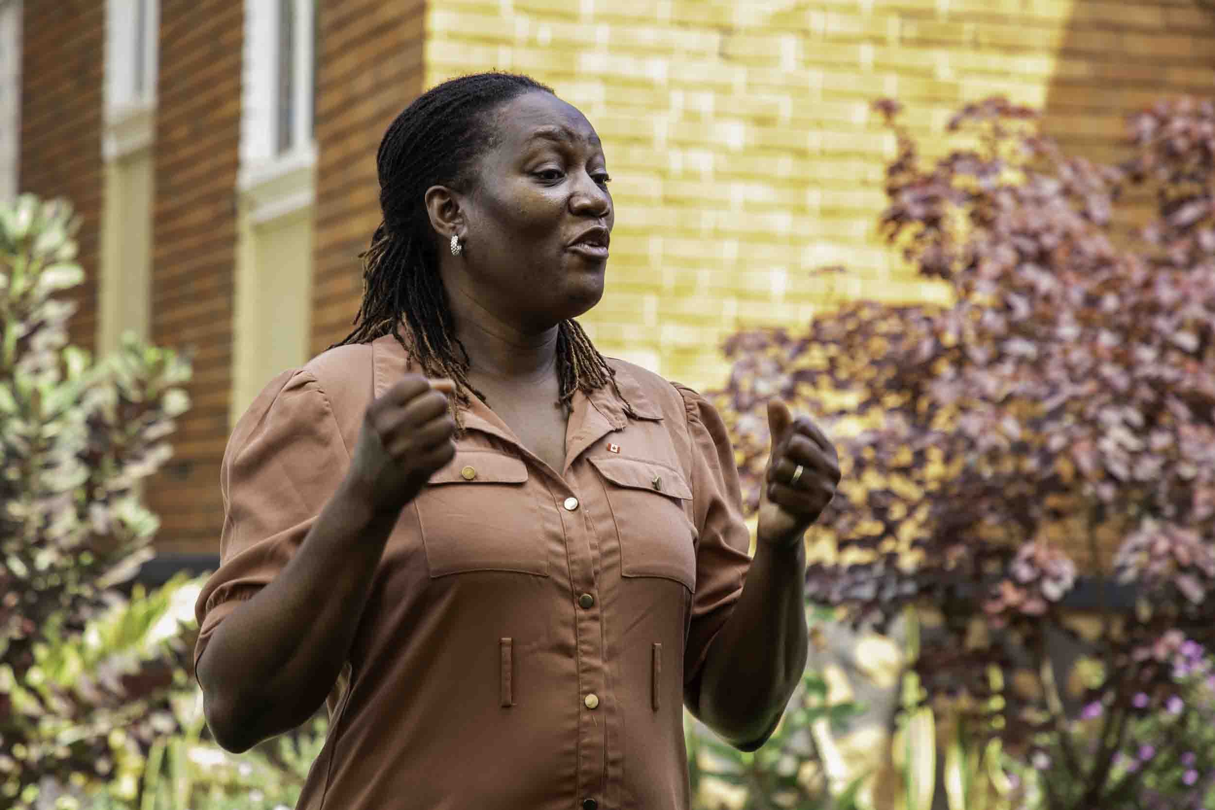 A woman with dark skin and dreadlocks wearing a brown shirt, speaking outdoors near a brick building with colorful plants in the background.