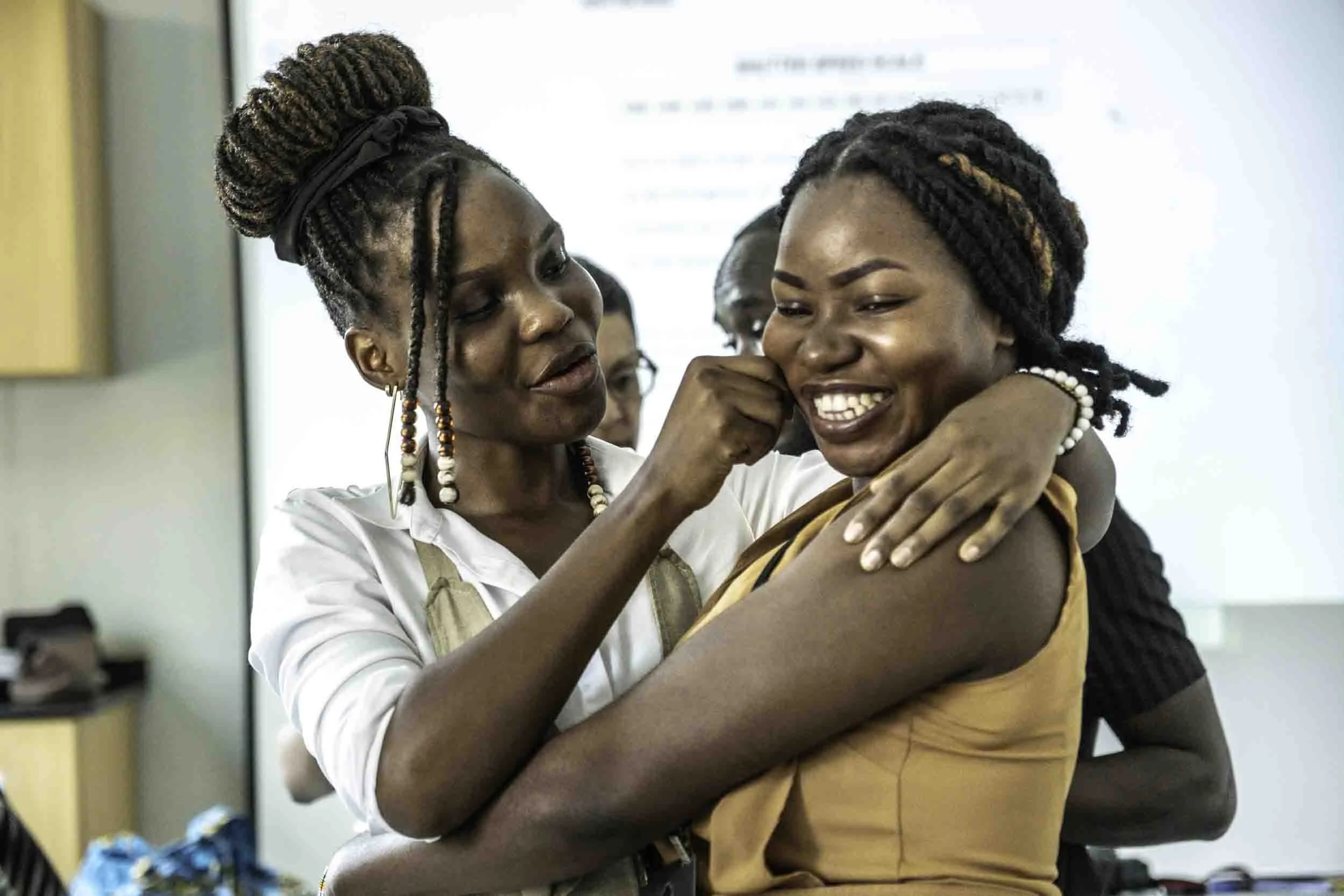 Two women share a joyful embrace in a classroom or office, one with her hair styled in a large updo and wearing earrings, the other with her hair in twists, both smiling.