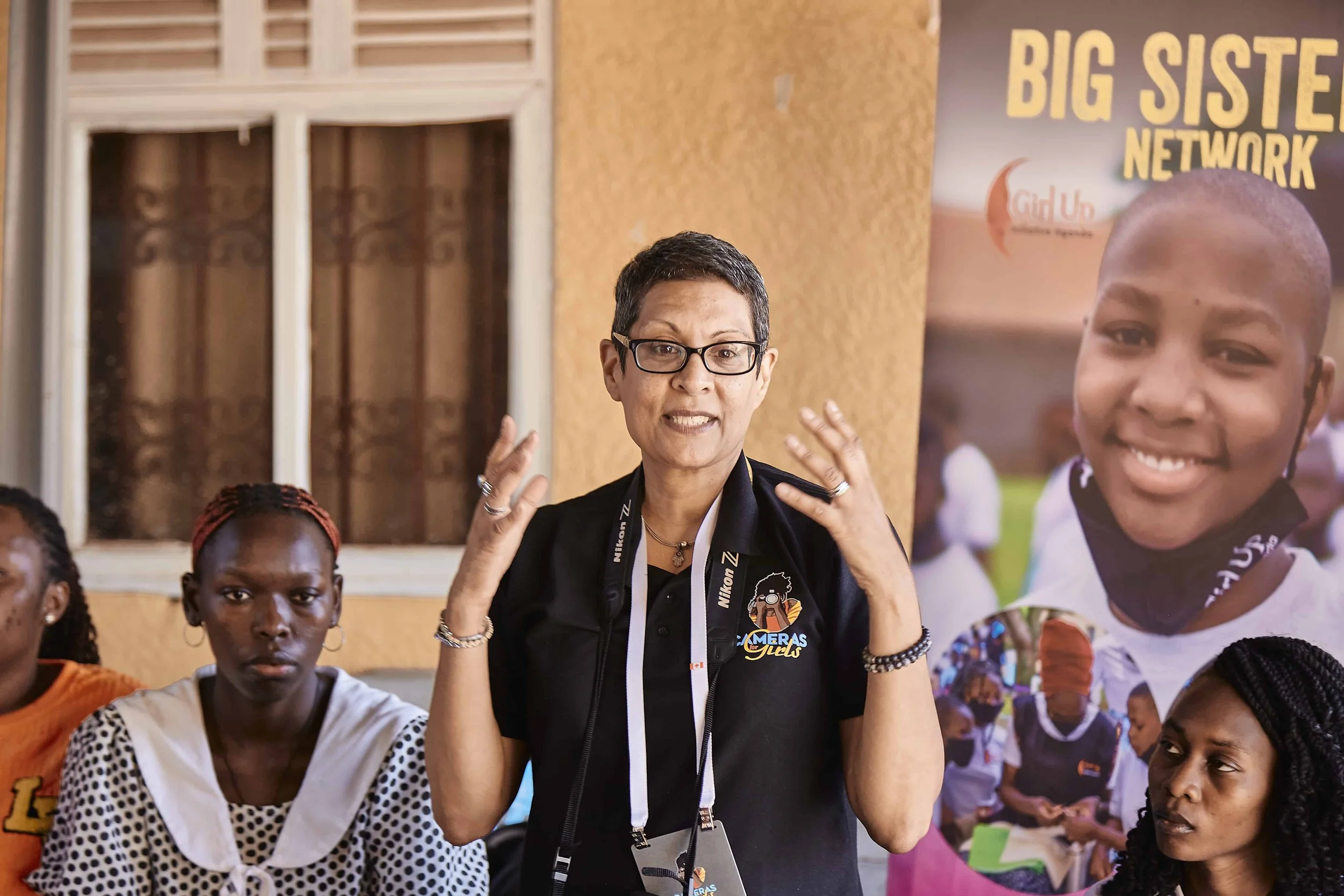 A woman with short hair and glasses speaking while surrounded by children, in front of a Big Sister Network sign and a photograph of a girl.