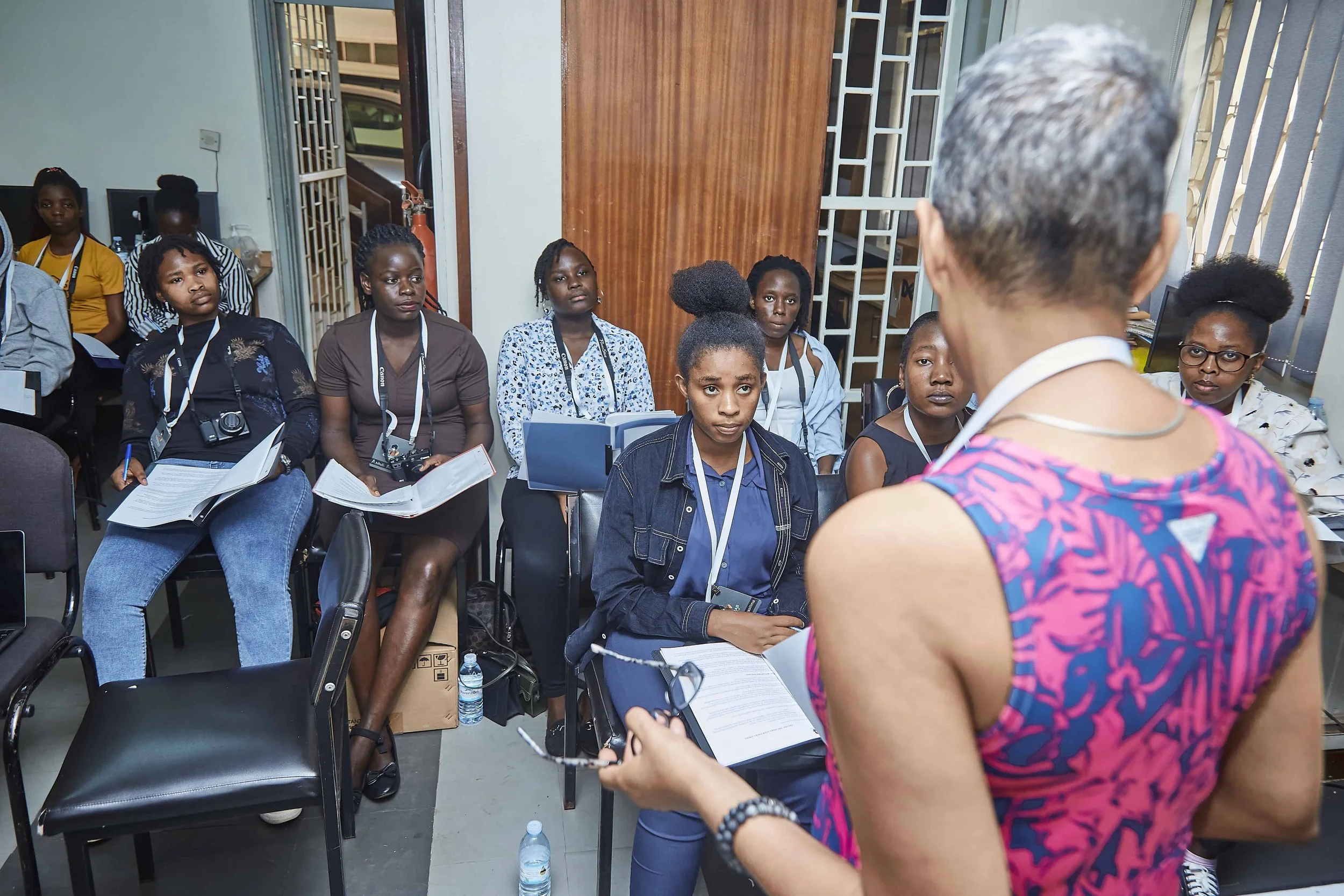 A woman in a pink and purple patterned sleeveless top is speaking to a group of mostly women seated in chairs, some holding notebooks and cameras. The audience members are paying attention, and some have cameras around their necks. The setting appear