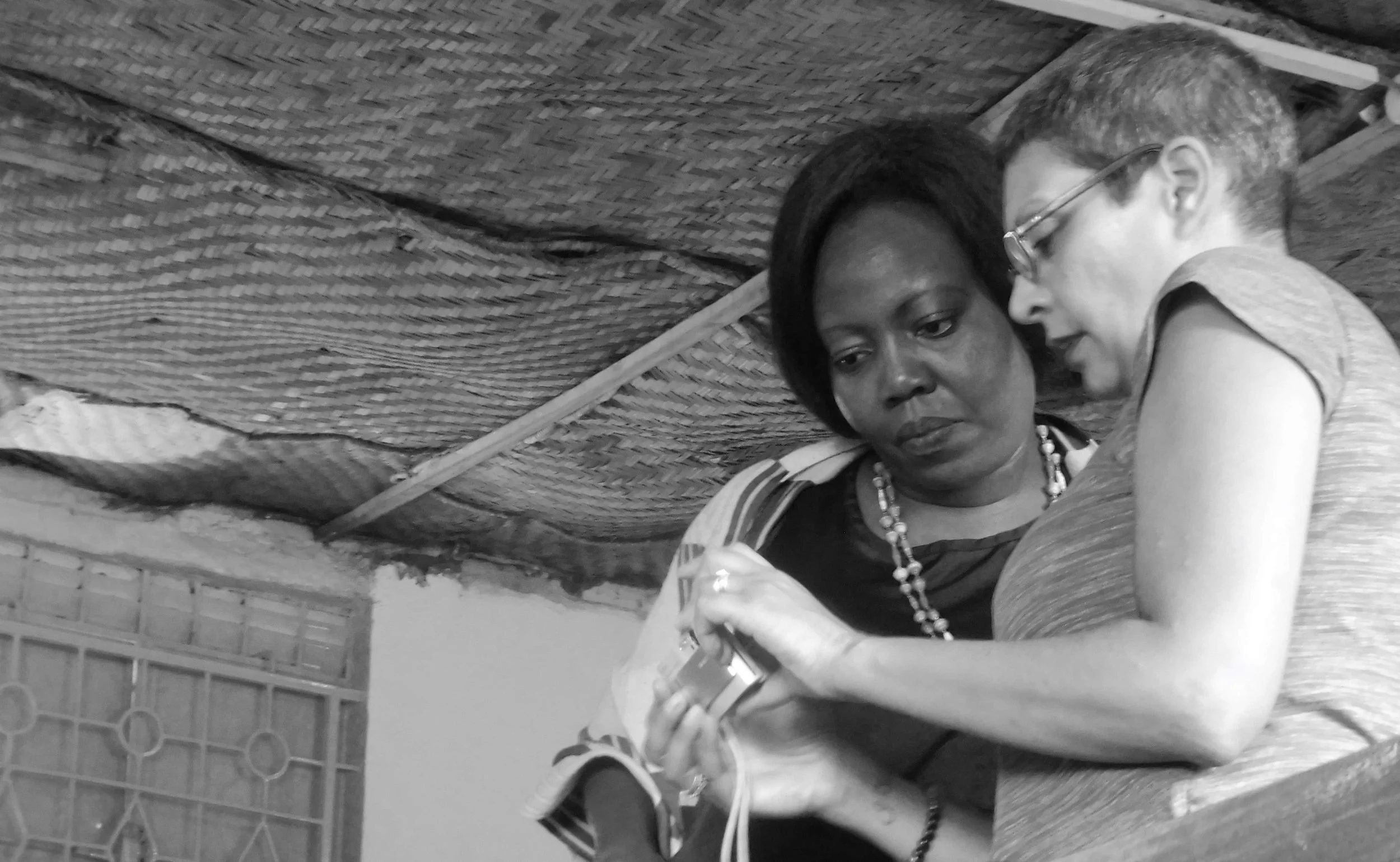 A woman and a young girl sitting close together under a thatched roof, looking at something in the woman's hands.
