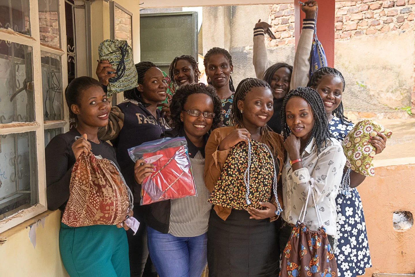 Group of ten women smiling and holding various items, standing in a brick-walled room with a window.