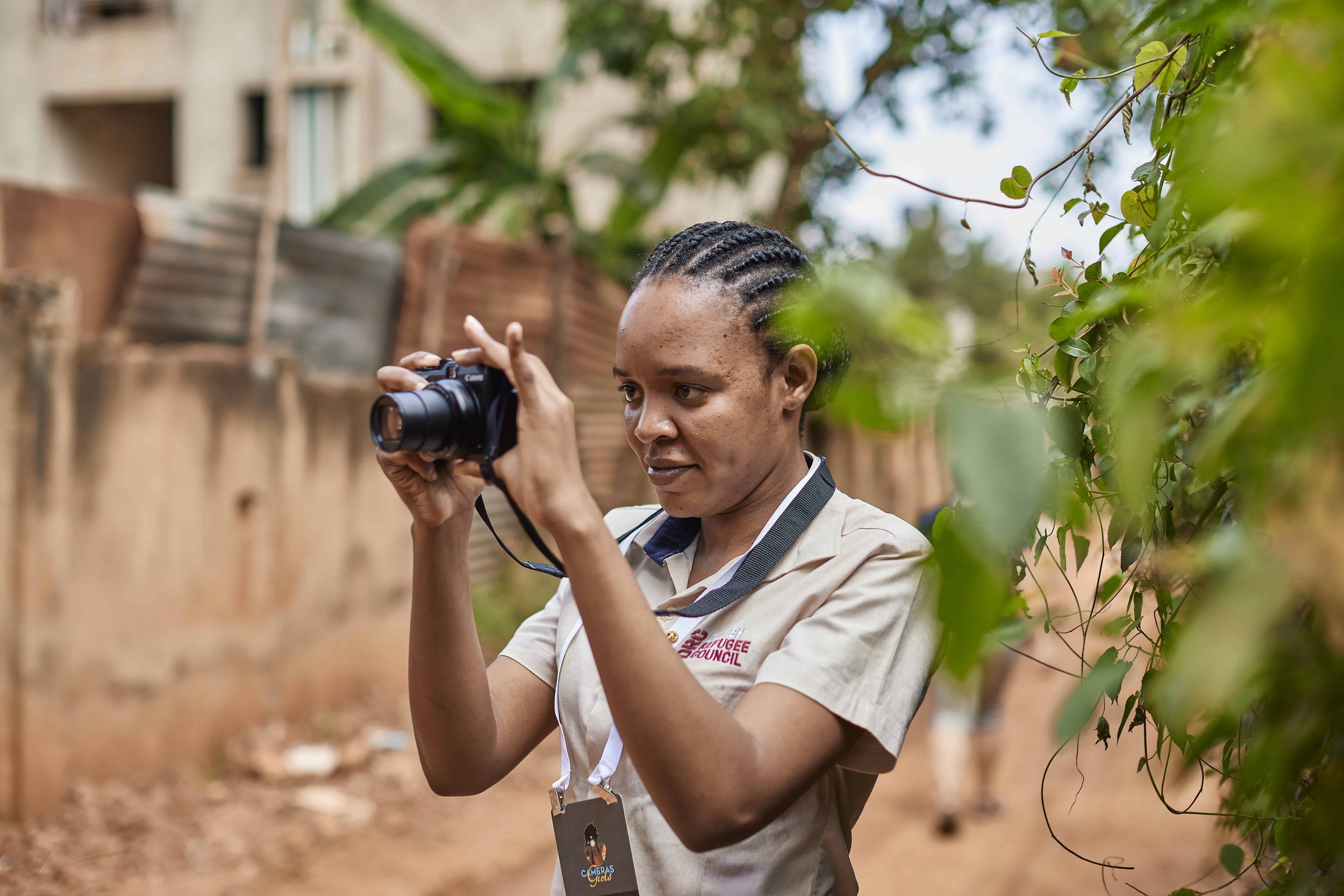 A woman with braided hair and beige shirt taking a photo with a camera outdoors among green plants and trees.