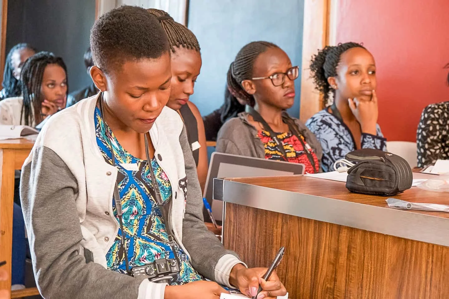 A group of women attending a lecture or seminar, sitting at desks, with some taking notes and one using a smartphone.