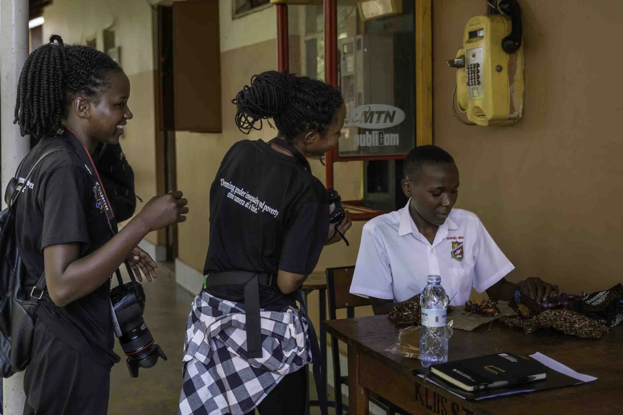 Three women and a young man gather around a table with items including water bottles, a laptop, and a cloth. The women are wearing black shirts, one with text on the back, and the young man is dressed in a white shirt. One woman is seated, and the ot