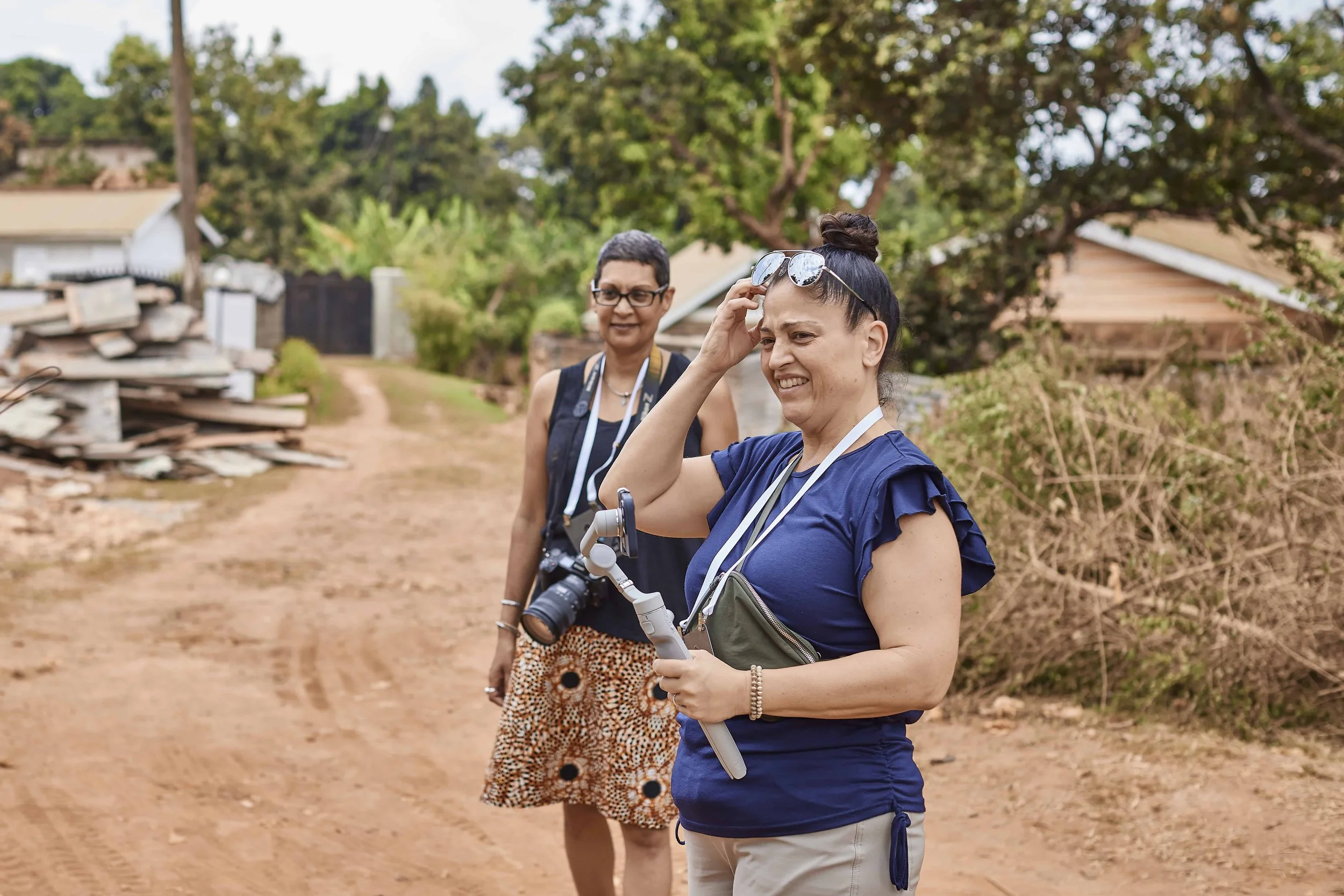 Two women outdoors on a dirt path, one adjusting sunglasses and the other with a camera around her neck, trees and a house in the background.