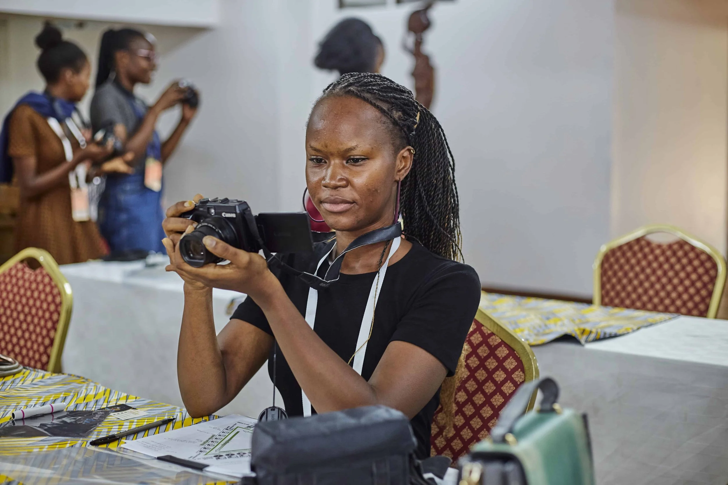 A woman with braided hair and hoop earrings is looking intently at a camera she is holding, seated at a table covered with a yellow and white patterned tablecloth inside a conference room.