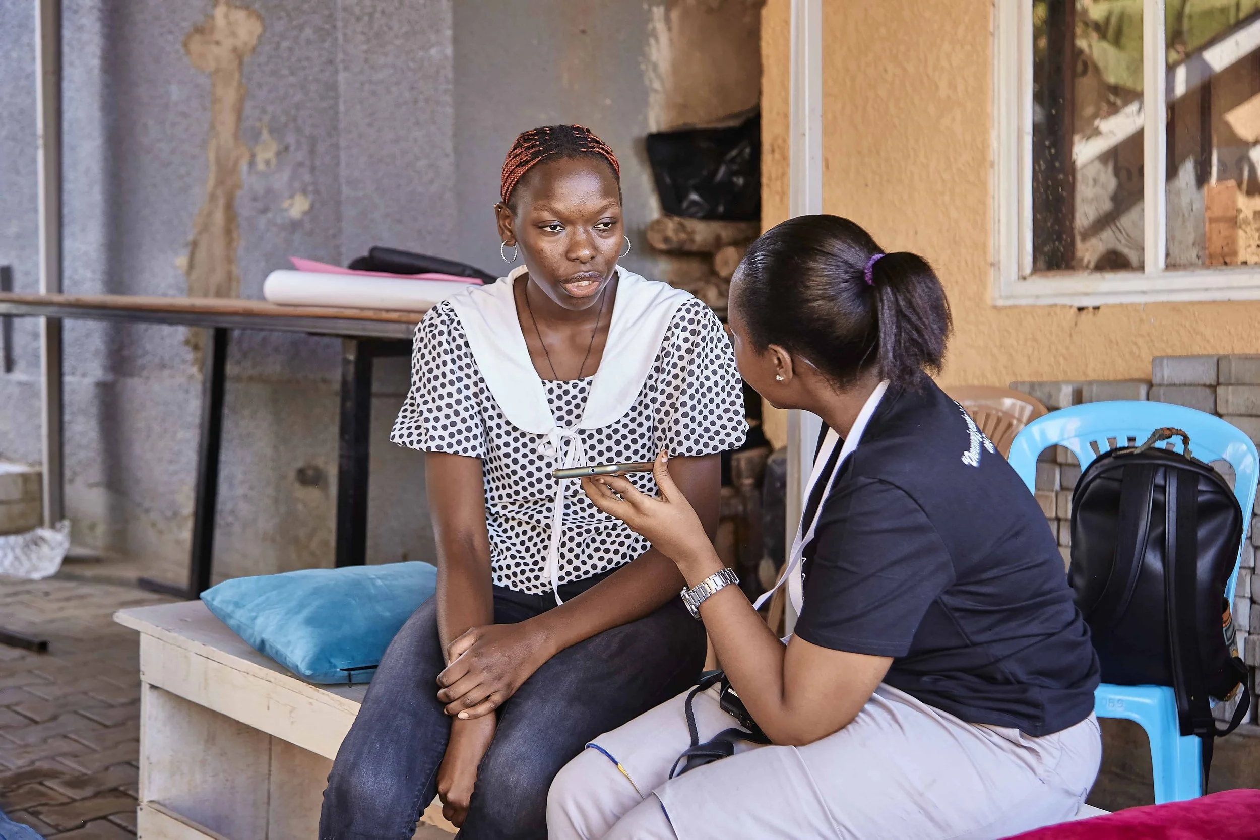 Two women sit on a bench, engaged in conversation; one woman is holding her phone to her ear, while the other looks attentively at her.