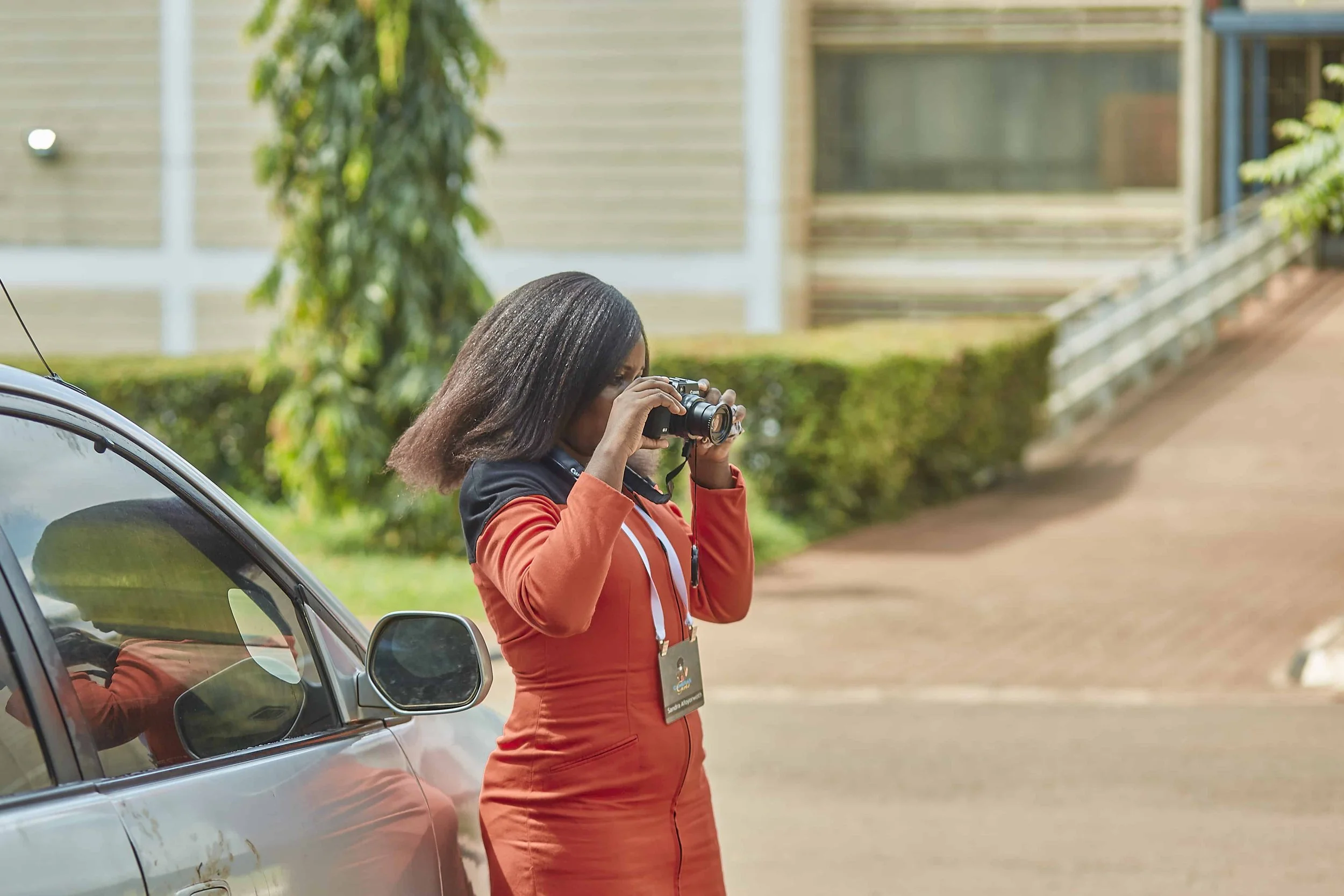 A woman with long dark hair taking a picture with a camera while standing next to a parked car outdoors.