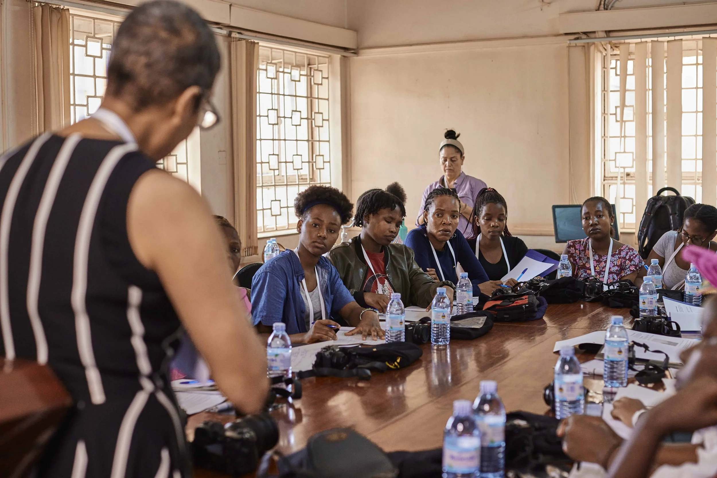 A group of women sitting at a long conference table in a bright room, listening to a woman standing at the front. The table has water bottles, papers, and electronic devices.