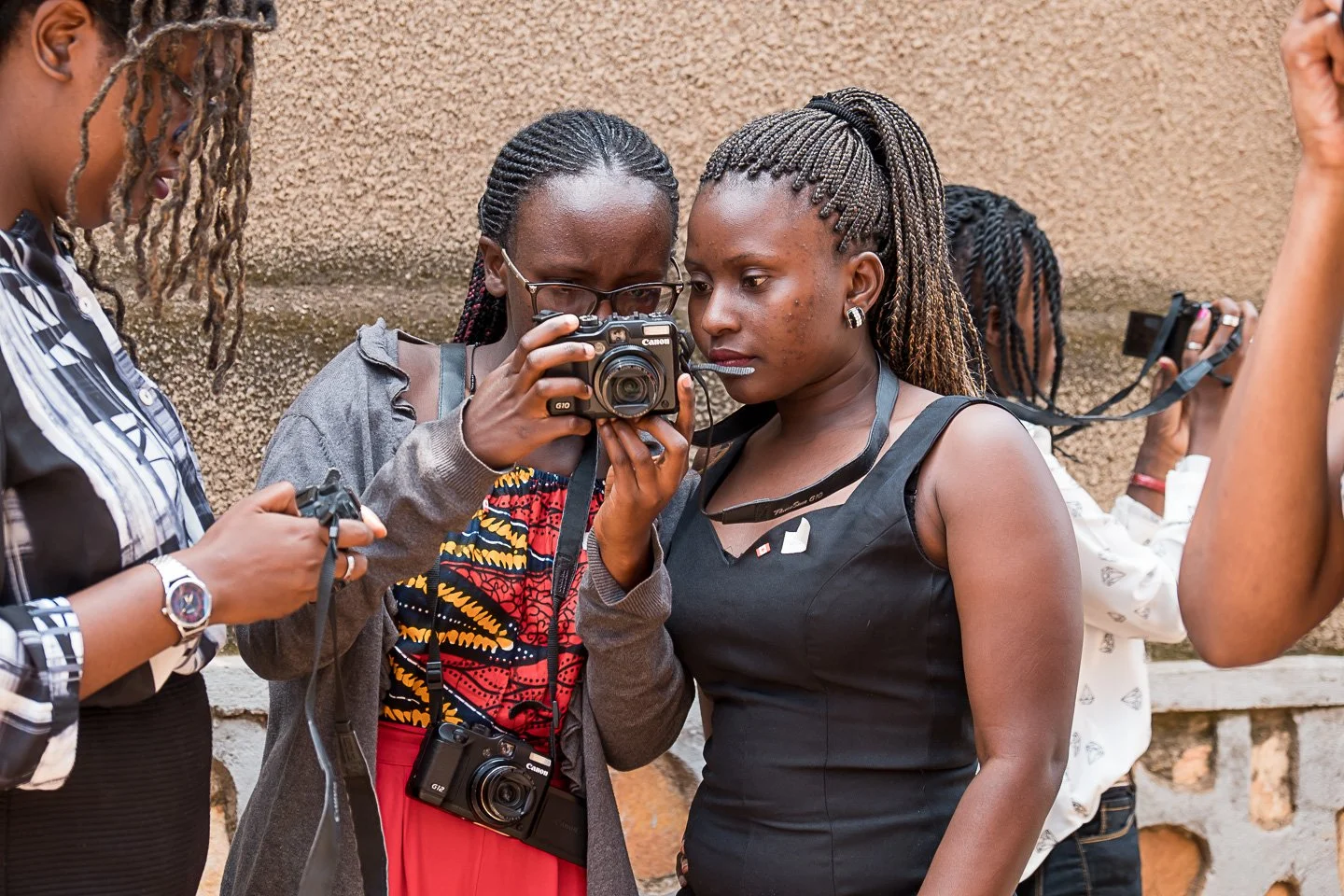 Three women are looking at a camera that one of them is holding, while another woman takes a photo of their scene. They are standing against a textured wall.