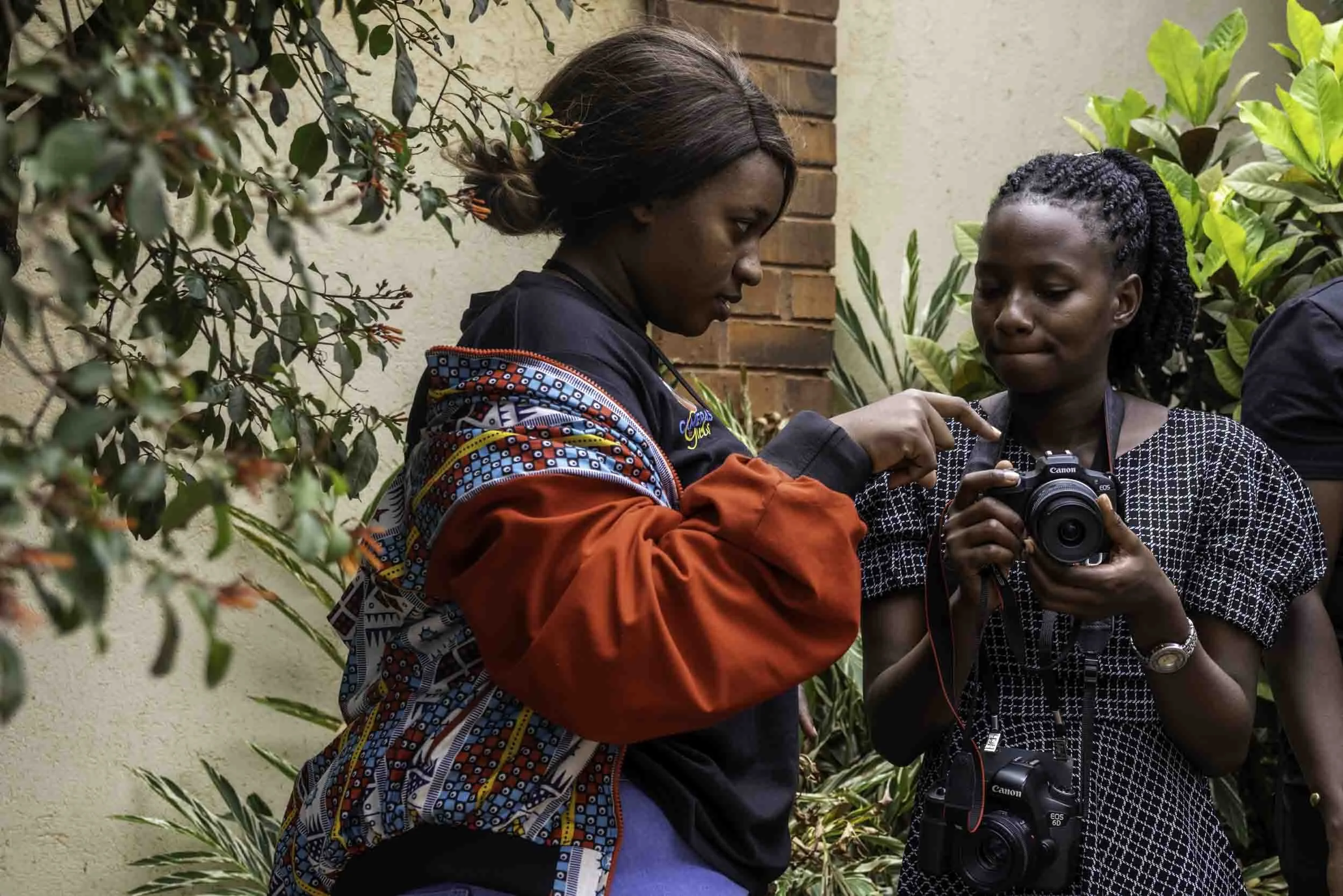 Two women, one with braided hair, are looking at a camera held by the other woman. They appear to be outdoors near a brick wall and green plants.