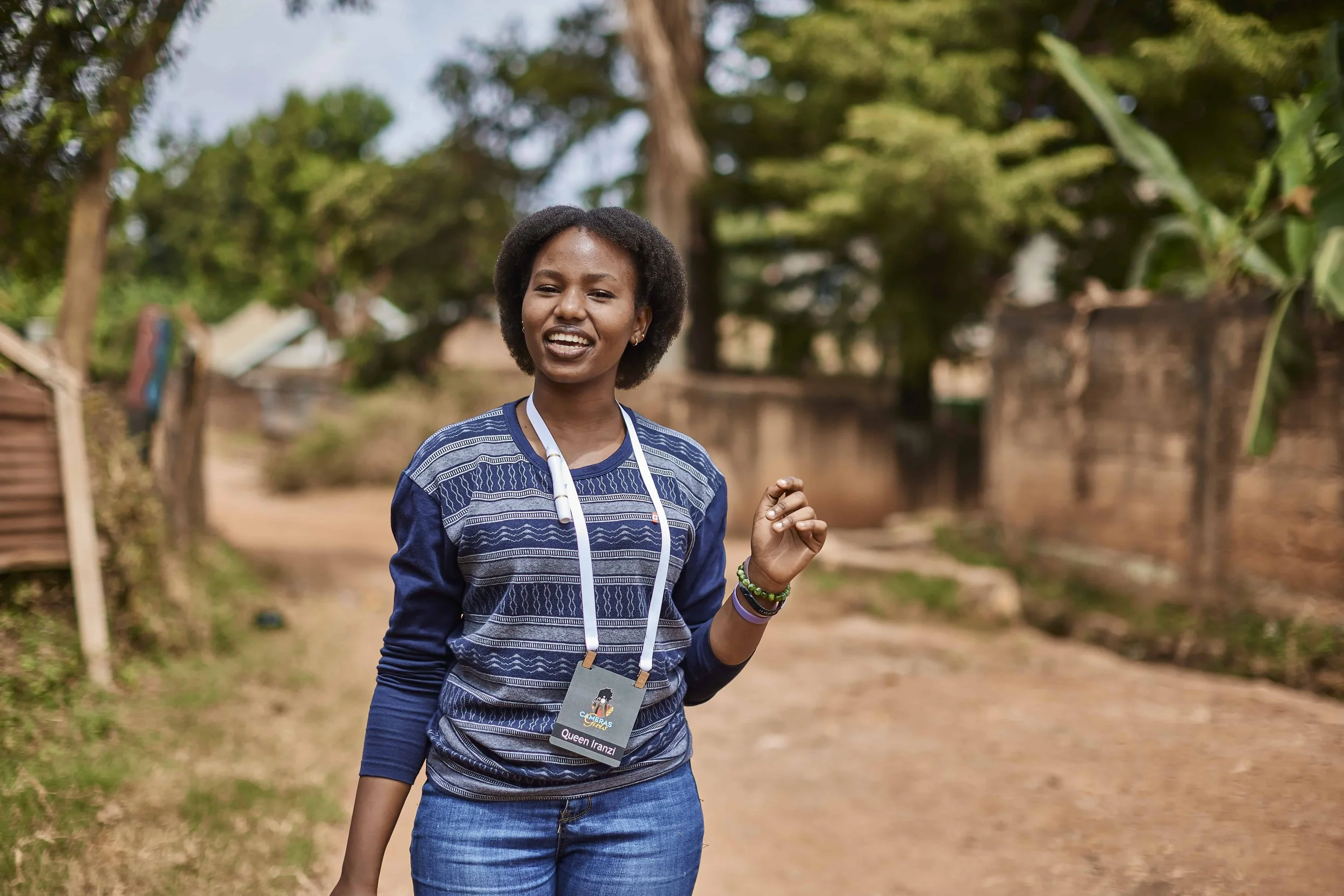 A woman with a name tag that reads Queen Iranzi, smiling outdoors with trees and a dirt path in the background.