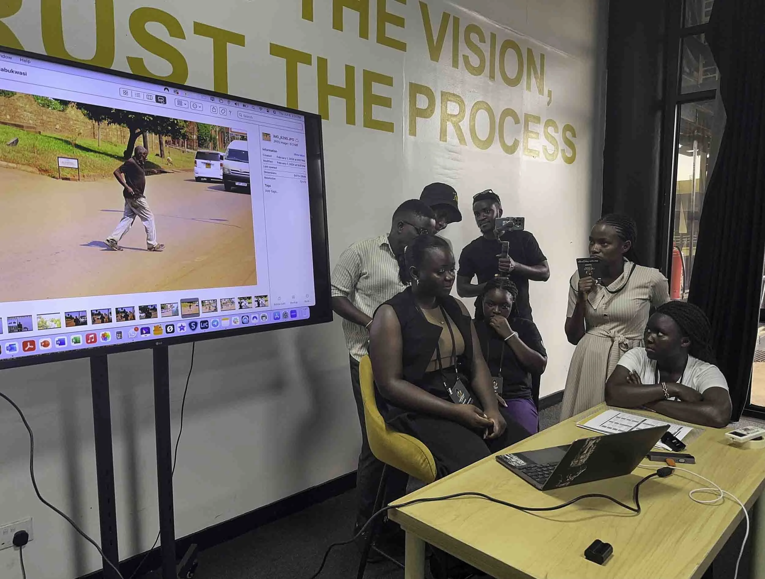 People gathered in a room with a large monitor showing an image of a man walking, alongside a yellow chair, a laptop, and various people taking notes or observing, with a wall displaying large yellow text that reads 'THE VISION, JUST THE PROCESS.'
