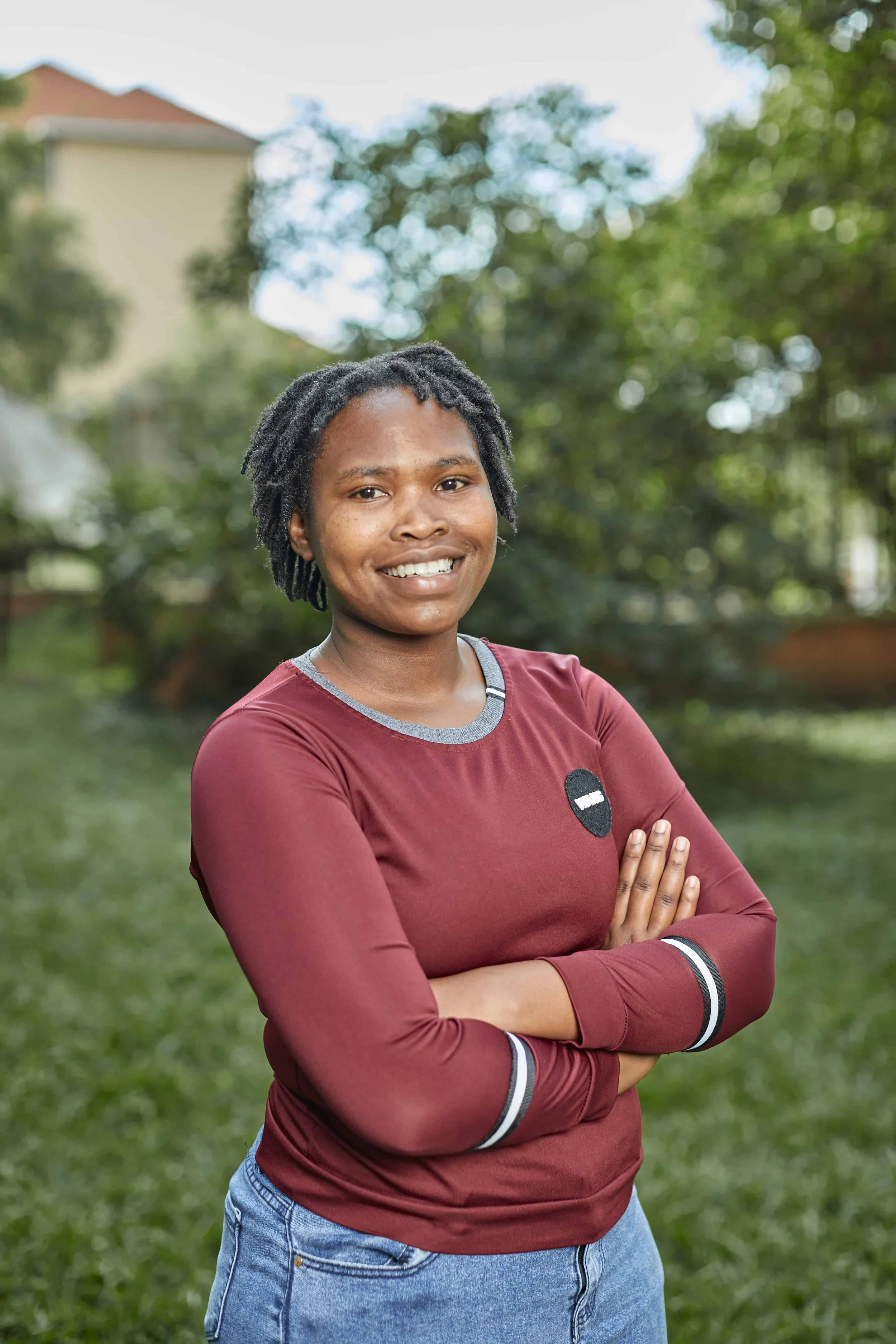 A woman with short, curly hair, smiling with arms crossed, wearing a maroon long-sleeve shirt and blue jeans, standing outdoors in a park with trees and a building in the background.