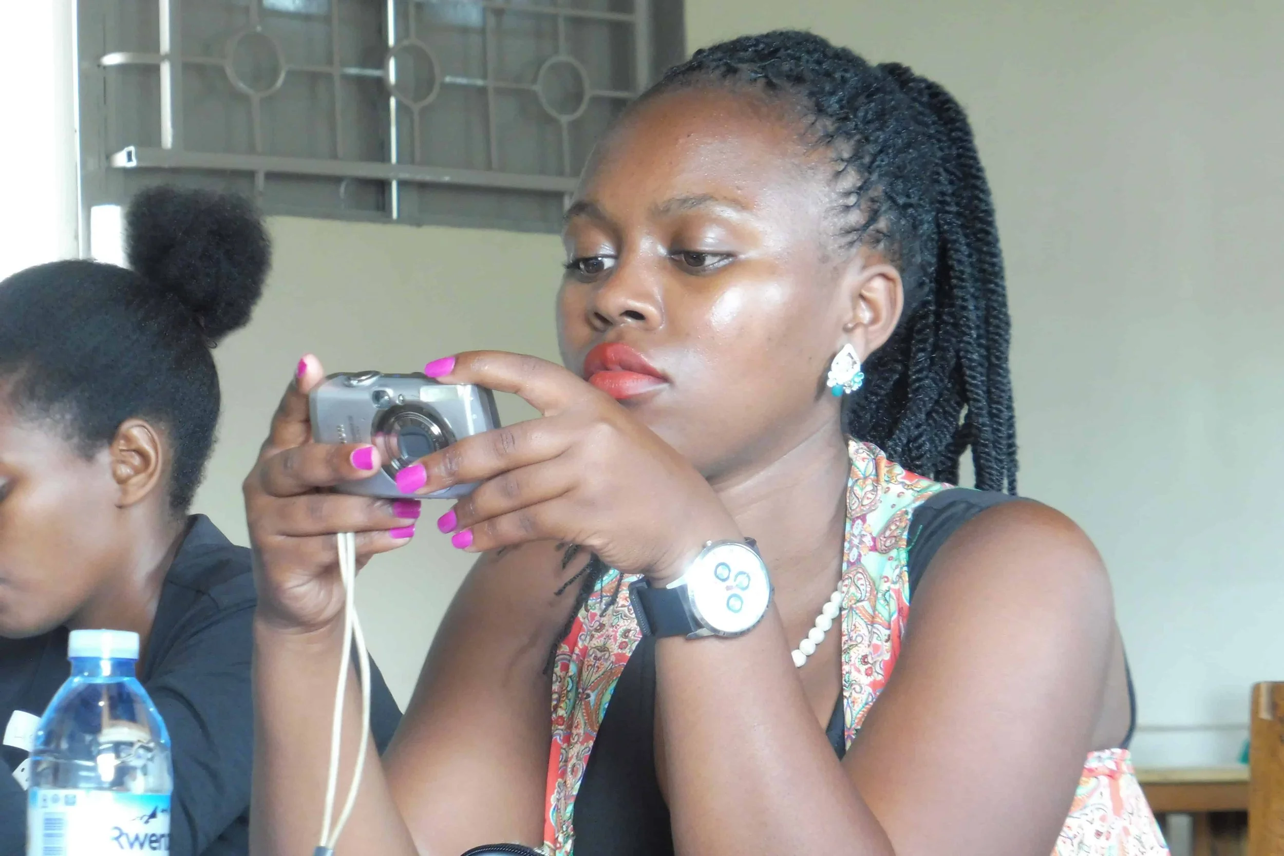 A woman with braided hair, wearing earrings, a watch, and a patterned dress, is looking at her phone. She is seated at a table with a water bottle and another person is partially visible beside her.